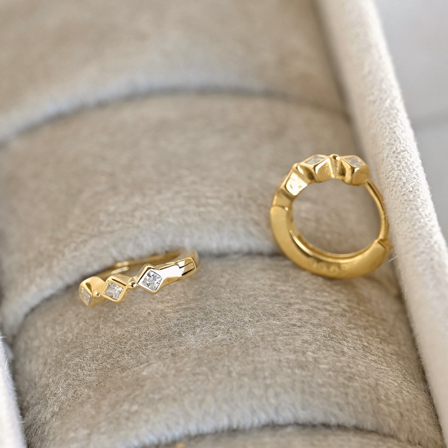 A pair of gold earrings with small geometric diamond-shaped facets displayed on a beige velvet jewelry box. 