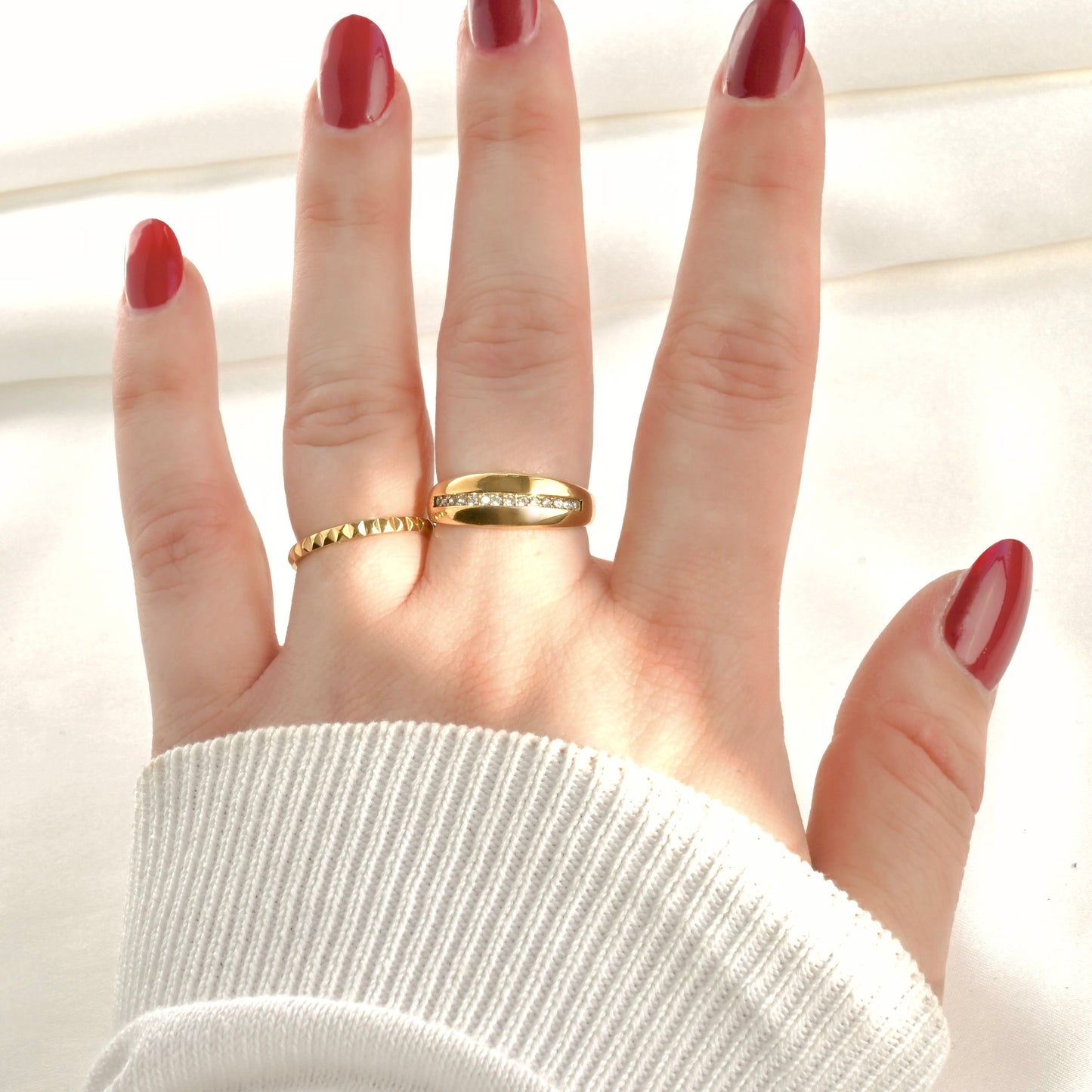 Hand wearing two gold rings, one thin pyramid ring and one cz line dome ring with red nail polish on a light background