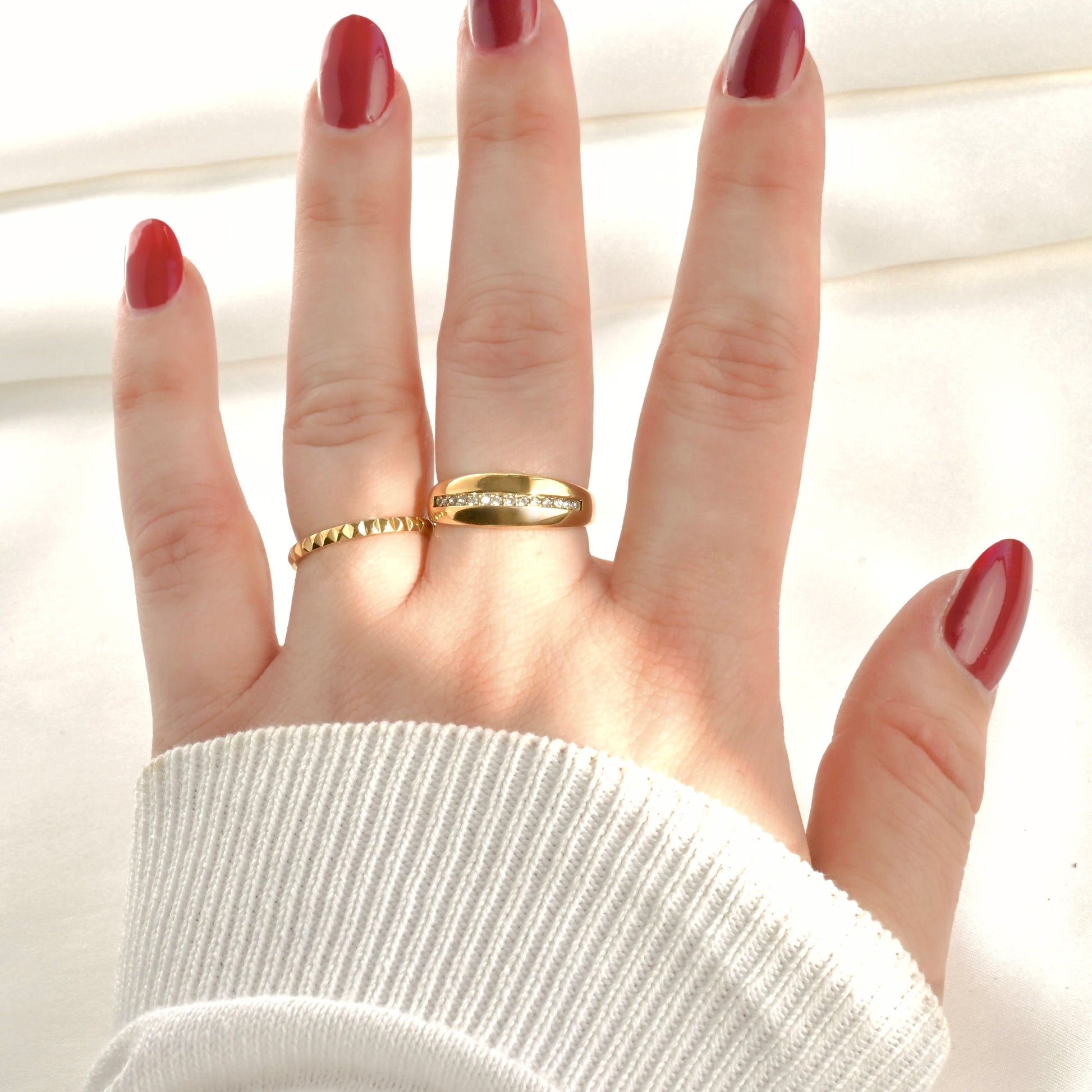 Hand wearing two gold rings, one thin pyramid ring and one cz line dome ring with red nail polish on a light background