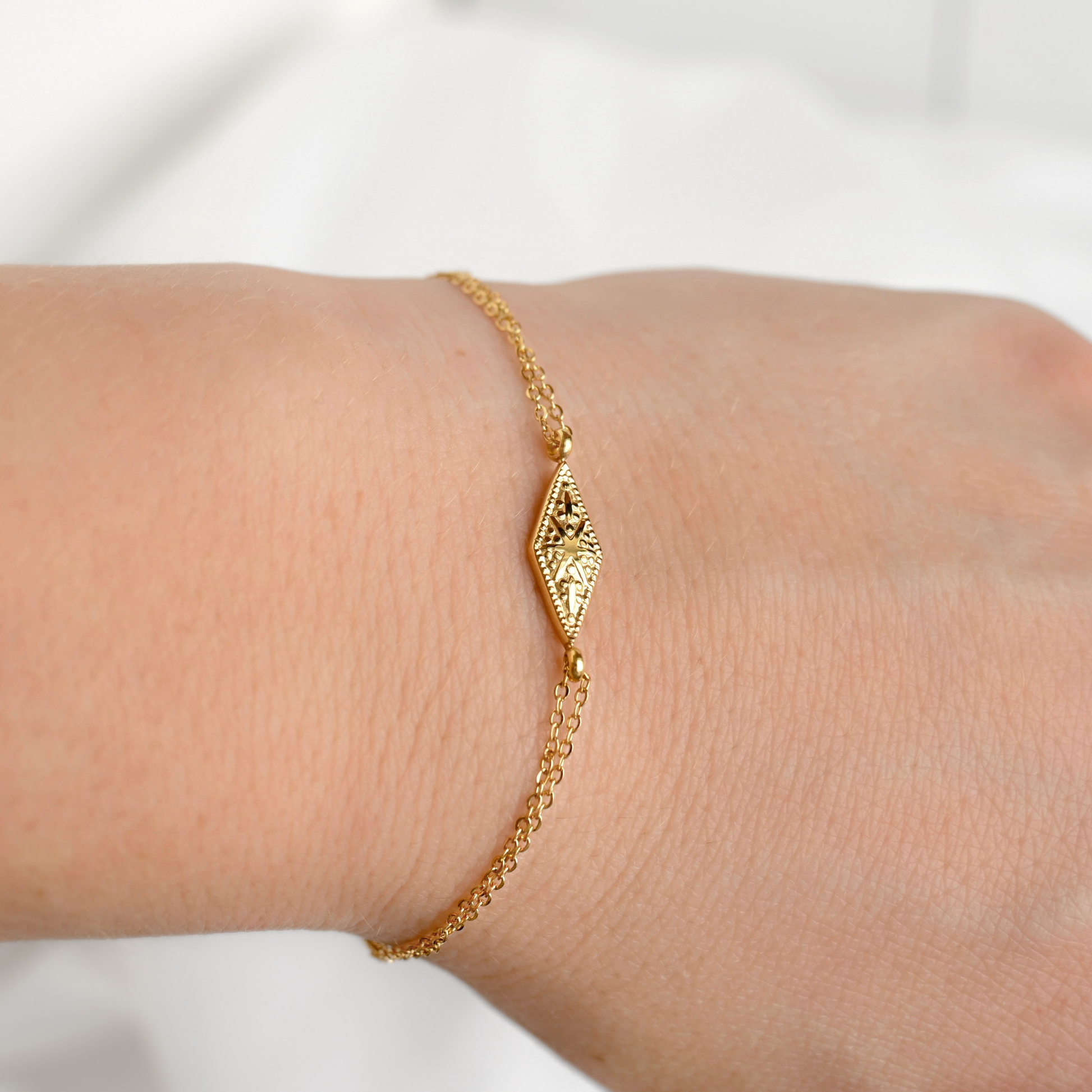 A delicate gold chain bracelet with a textured, diamond-shaped sunburst charm, shown worn on a person's wrist against a soft, light background.