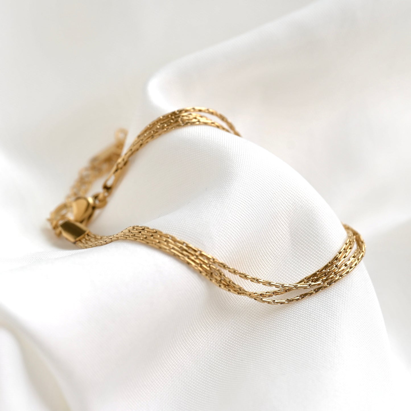 A delicate gold chain bracelet draped over soft white satin fabric.