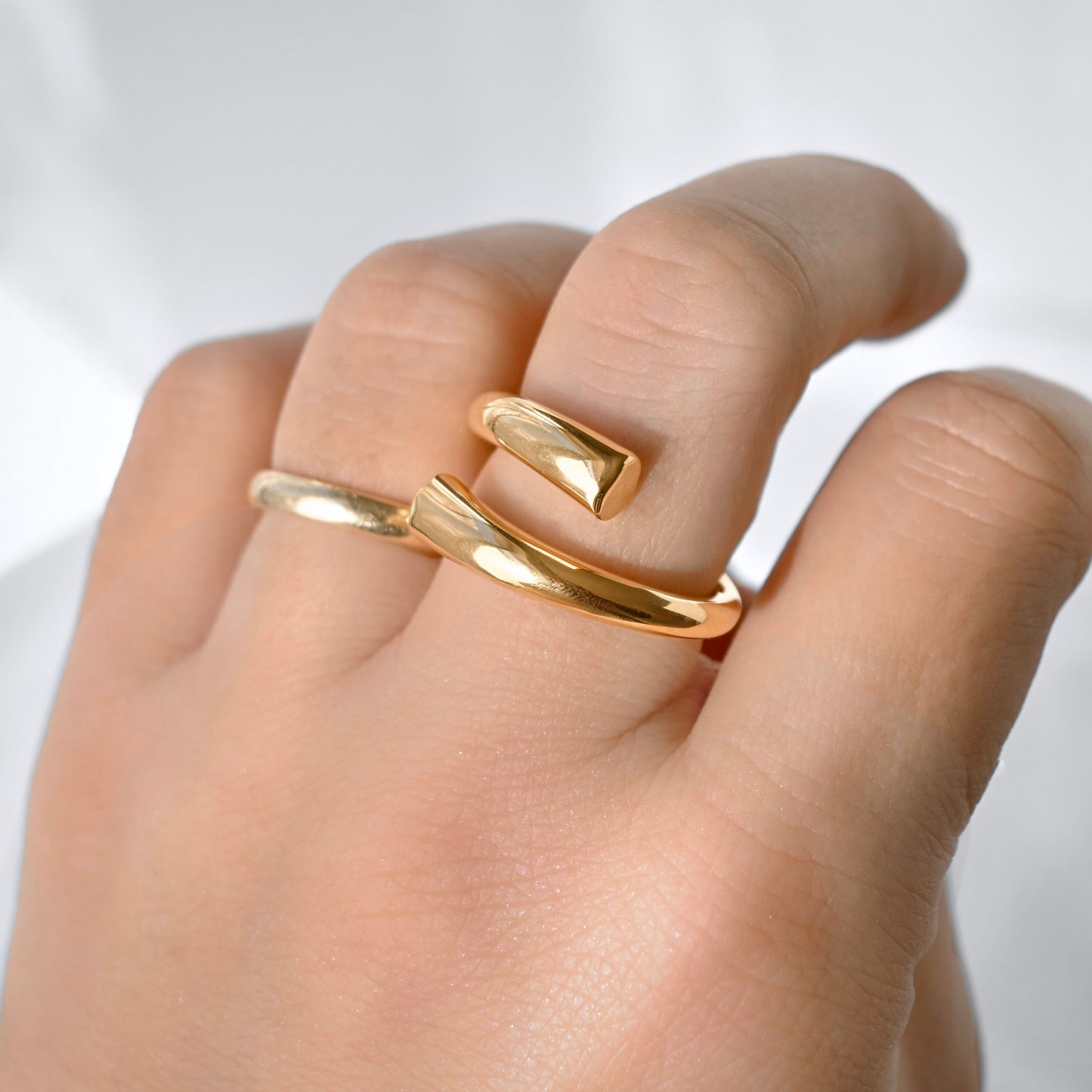 A close-up of a hand wearing a polished gold open wrap ring, showing the smooth metallic band in use on the fingers against a soft white background.