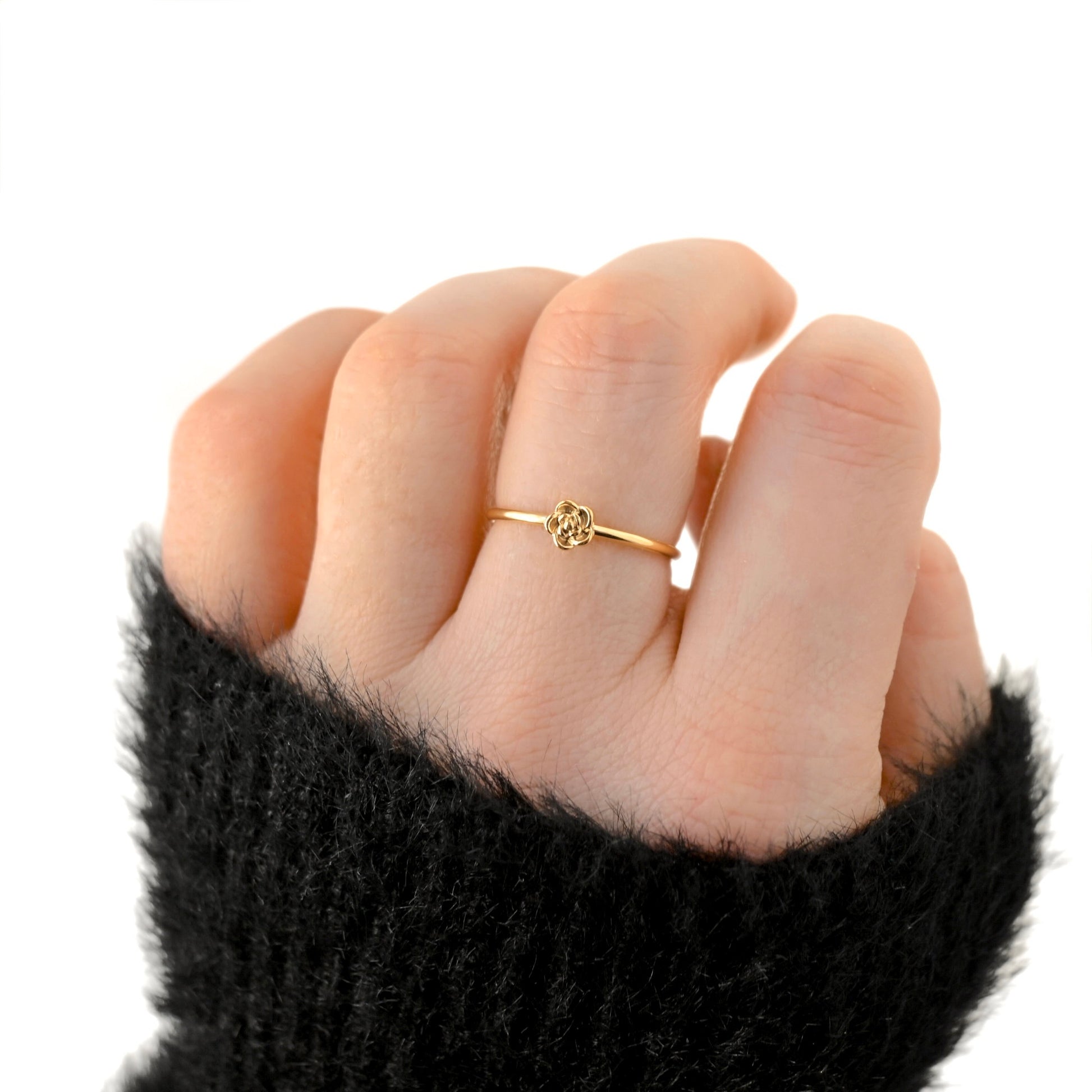 A delicate gold ring with a small floral or clover-like motif worn on a person's ring finger, shown against a plain white background. The hand is wearing a black fuzzy knit sleeve.