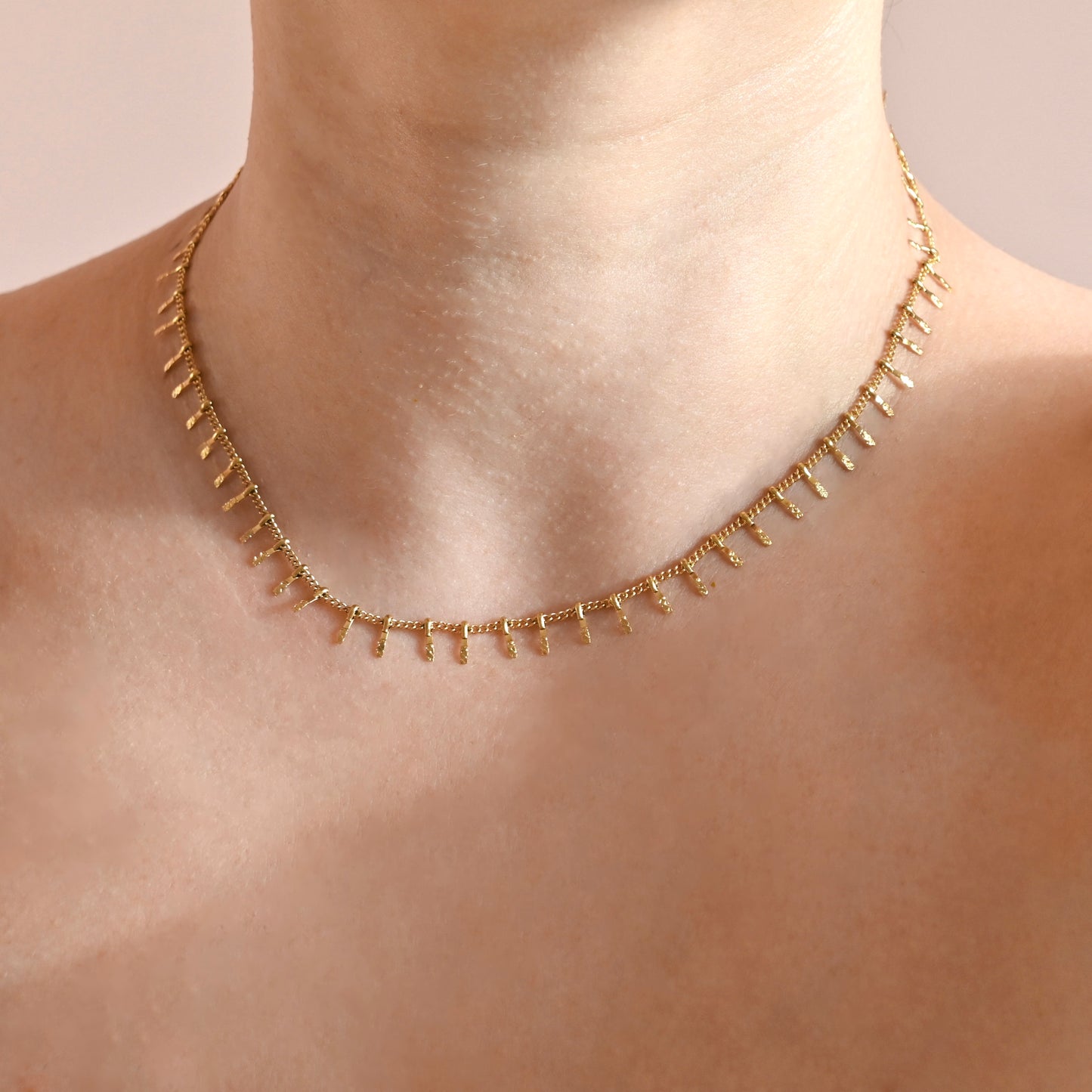 A delicate gold chain necklace with small dangling spikes, shown worn on a model's neck against a soft beige background.