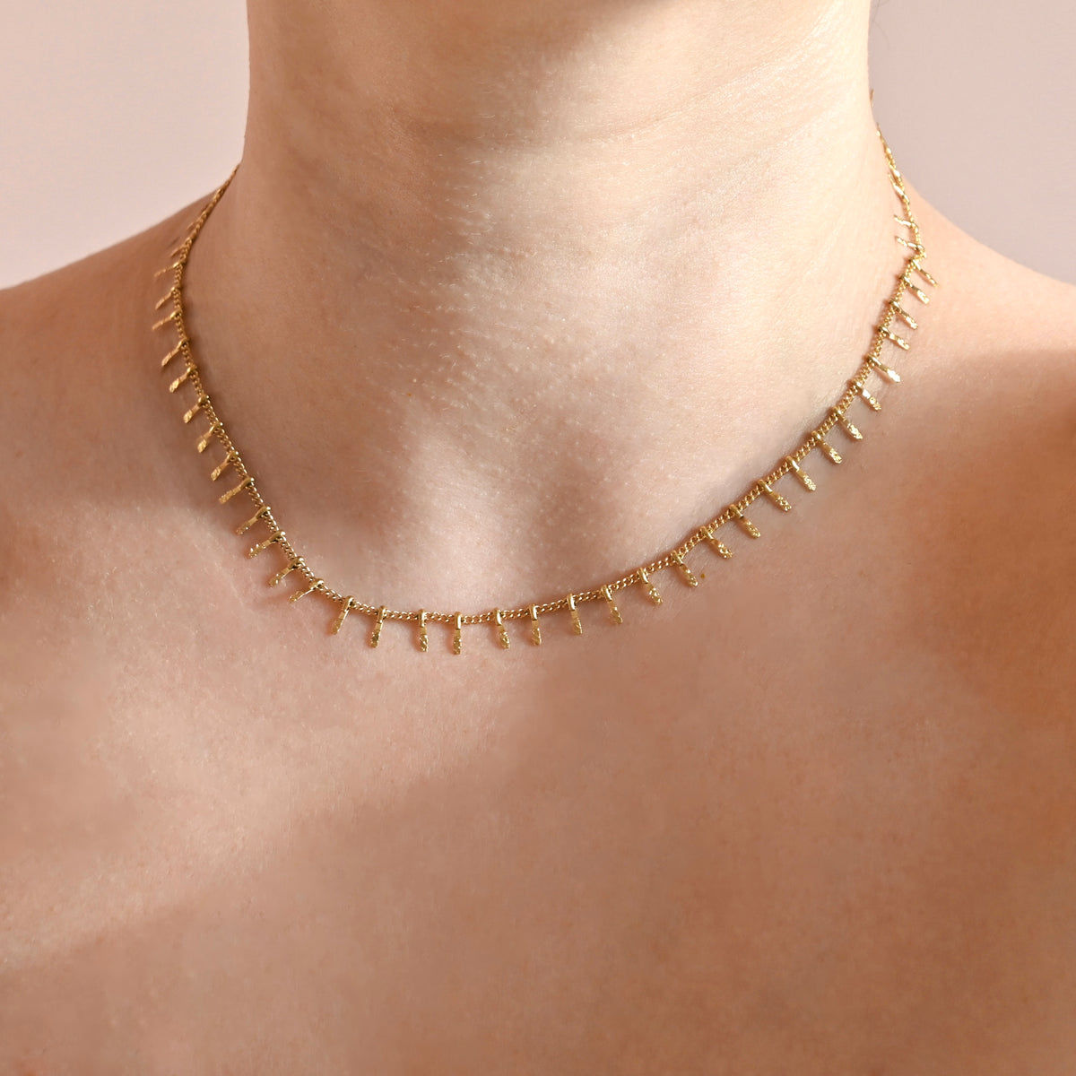 A delicate gold chain necklace with small dangling spikes, shown worn on a model's neck against a soft beige background.