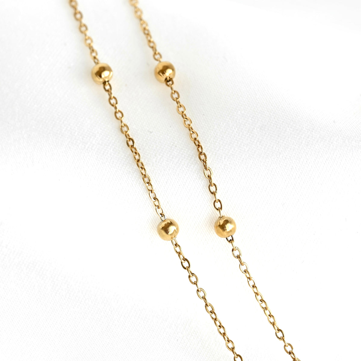 A delicate gold chain necklace featuring four evenly spaced, polished round gold beads, displayed on a soft white fabric background.
