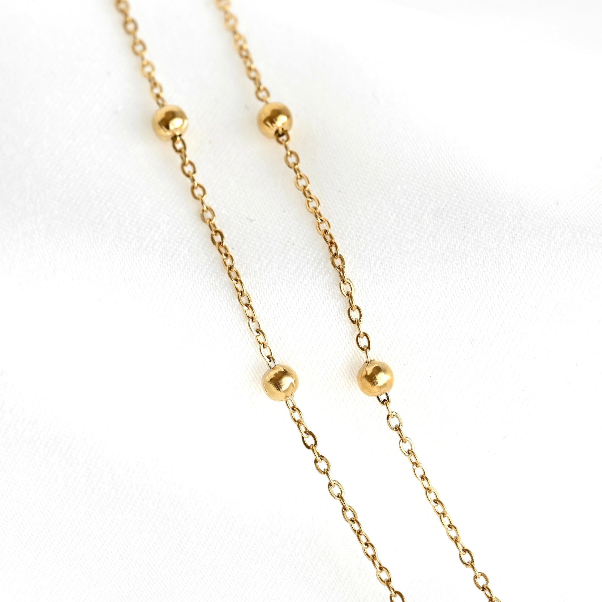 A delicate gold chain necklace featuring four evenly spaced, polished round gold beads, displayed on a soft white fabric background.