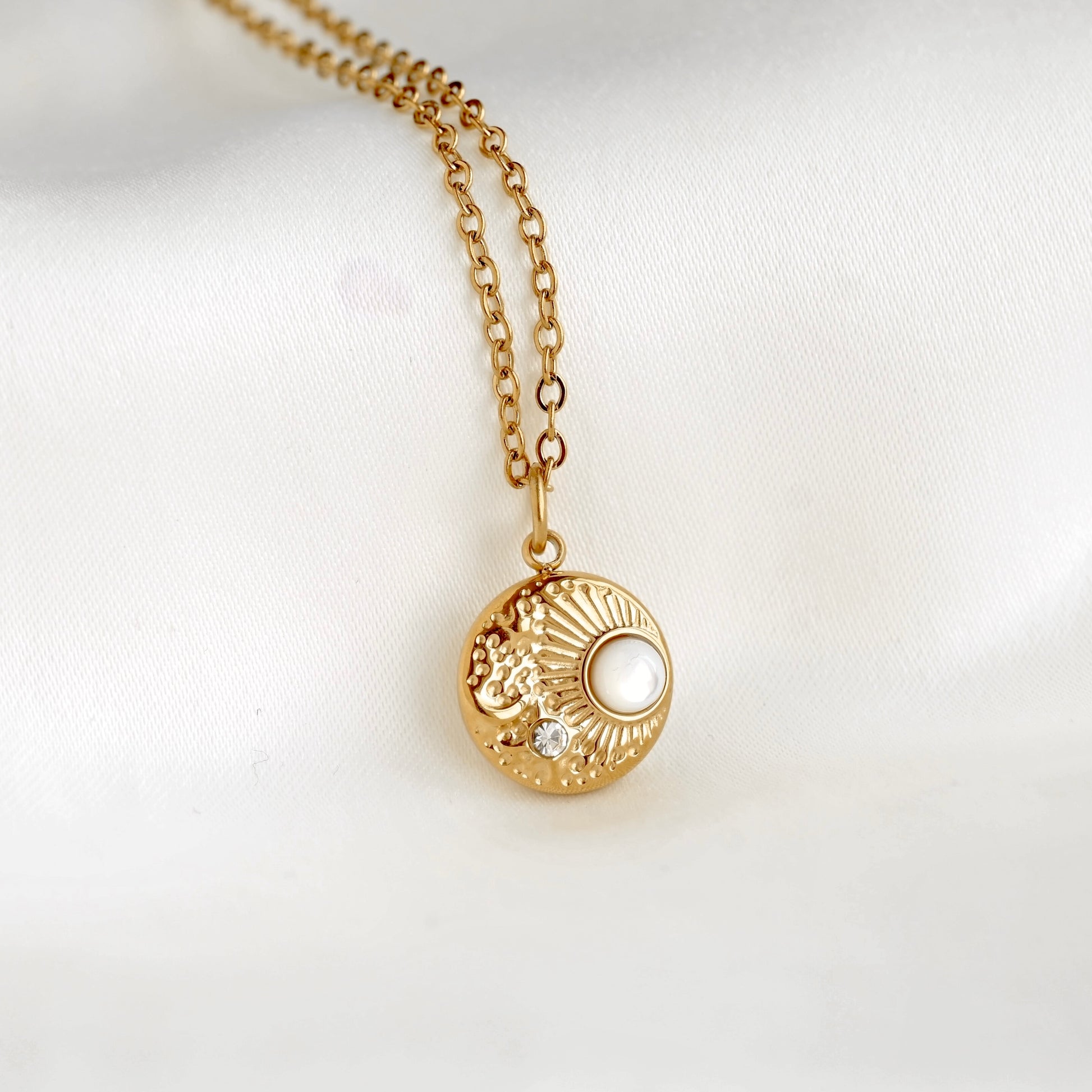 A gold pendant necklace featuring a textured sunburst moon design with a white pearl and a small clear crystal, resting on a white satin fabric background.