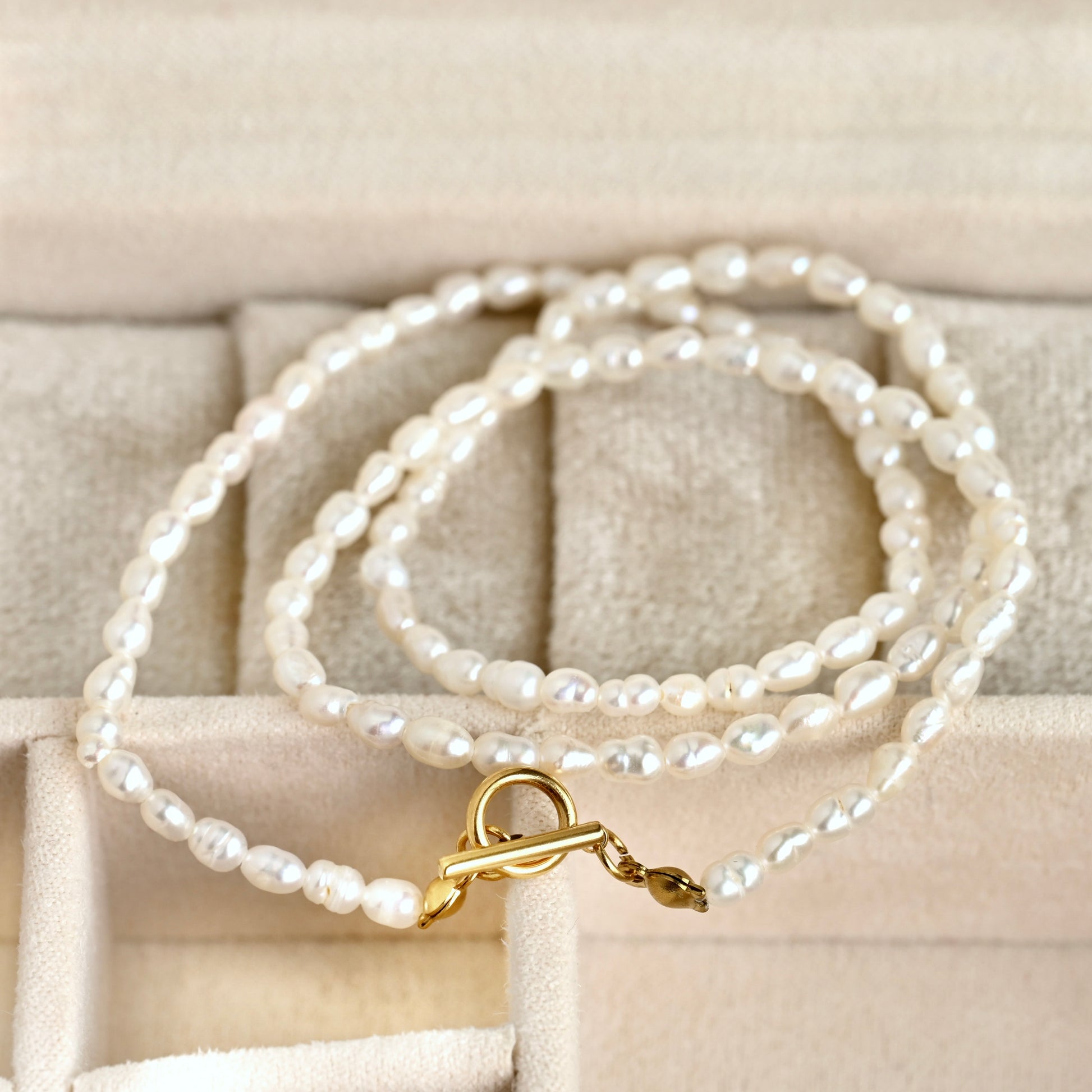 A strand of irregular white freshwater pearls with a gold toggle clasp, displayed on a beige jewelry box.