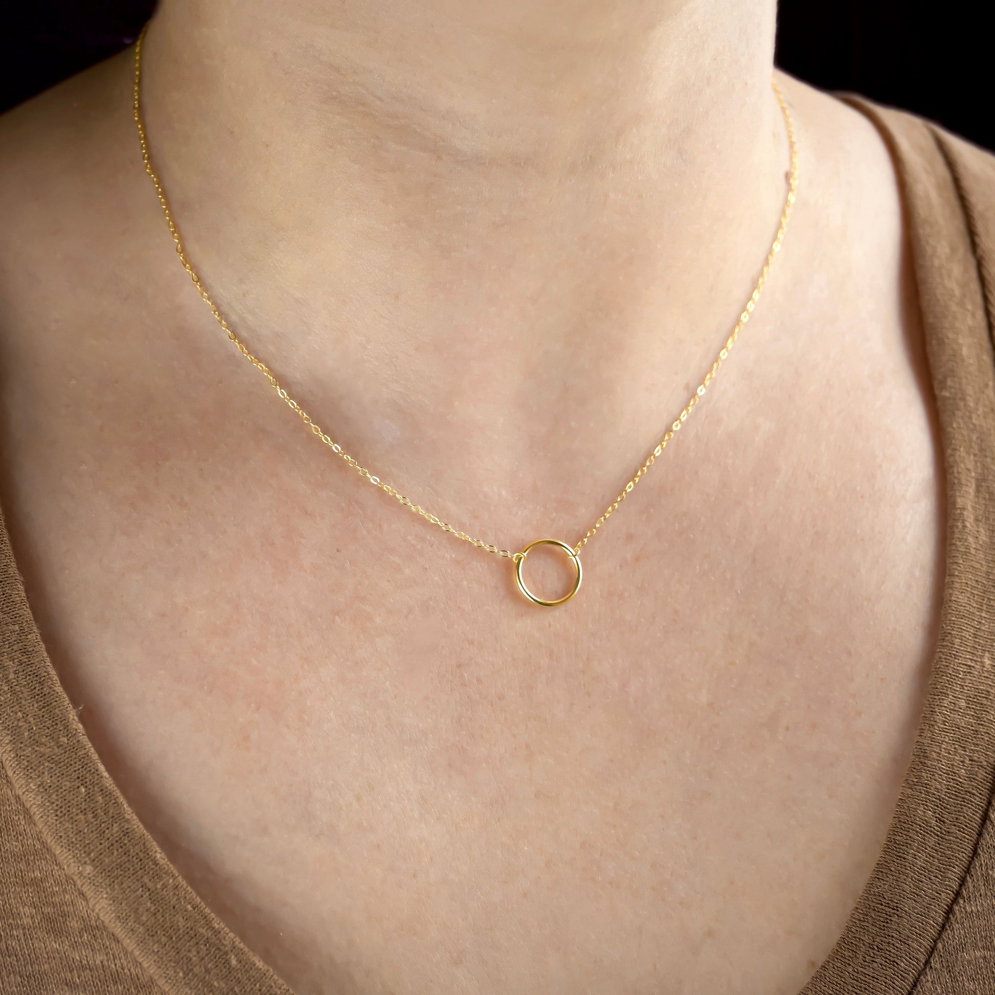 Gold necklace with a small ring pendant on a person wearing a brown top.