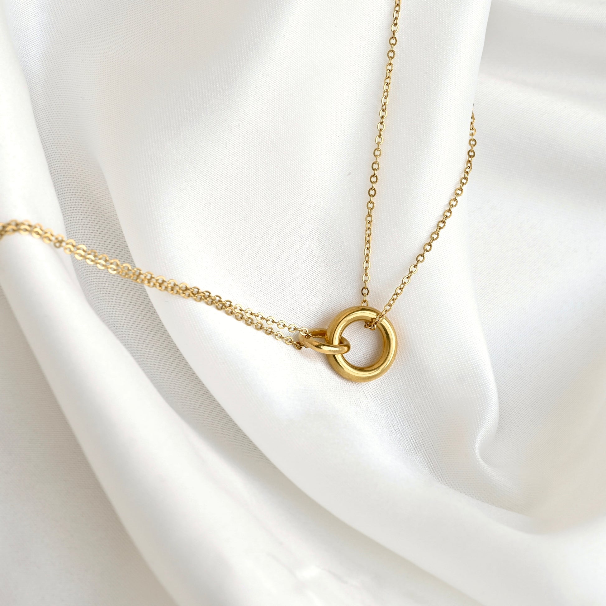 A delicate gold chain necklace with a minimalist interlocking double-ring pendant, displayed on draped white satin fabric.