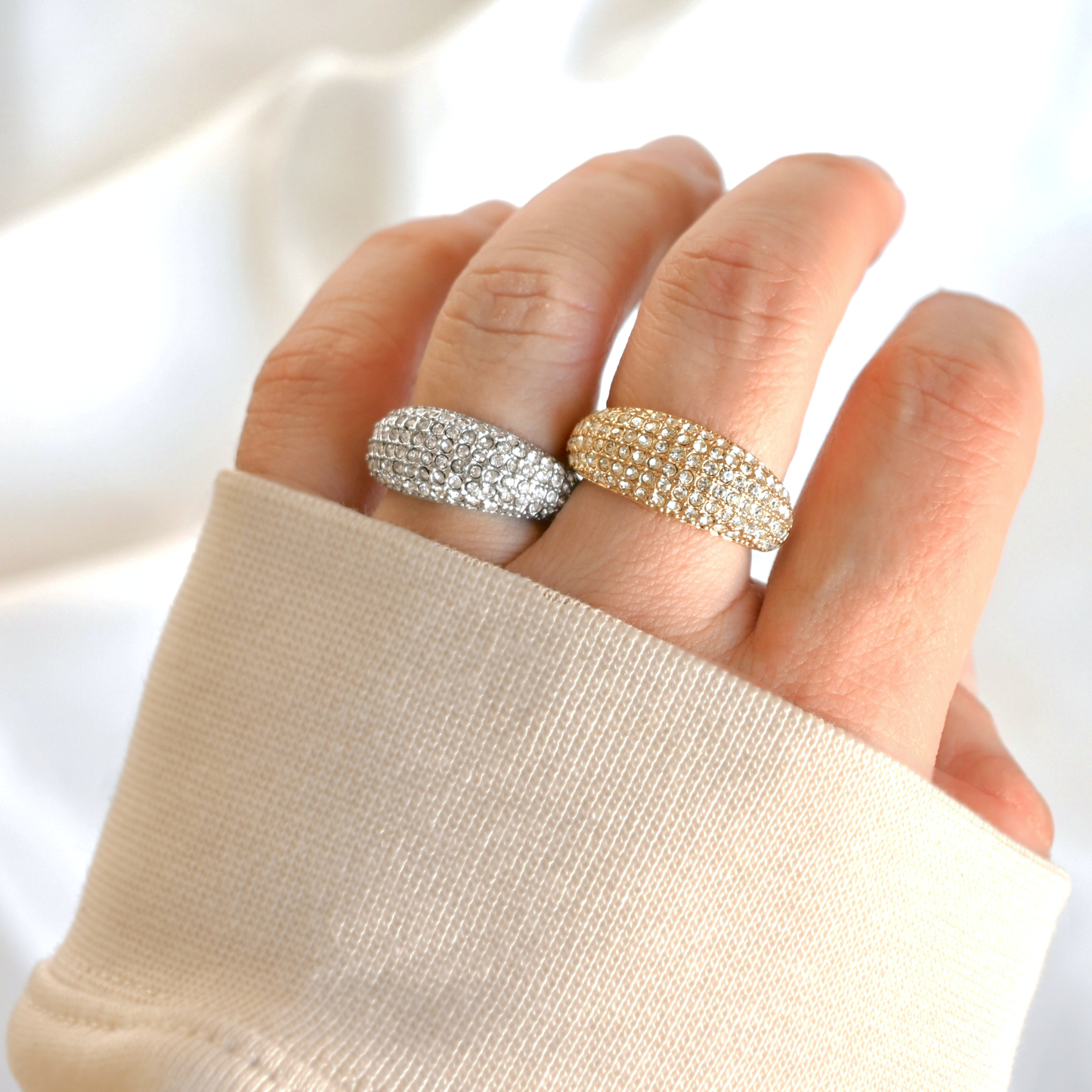 Hand wearing two chunky pave diamond rings, one gold and one silver, on a plain background