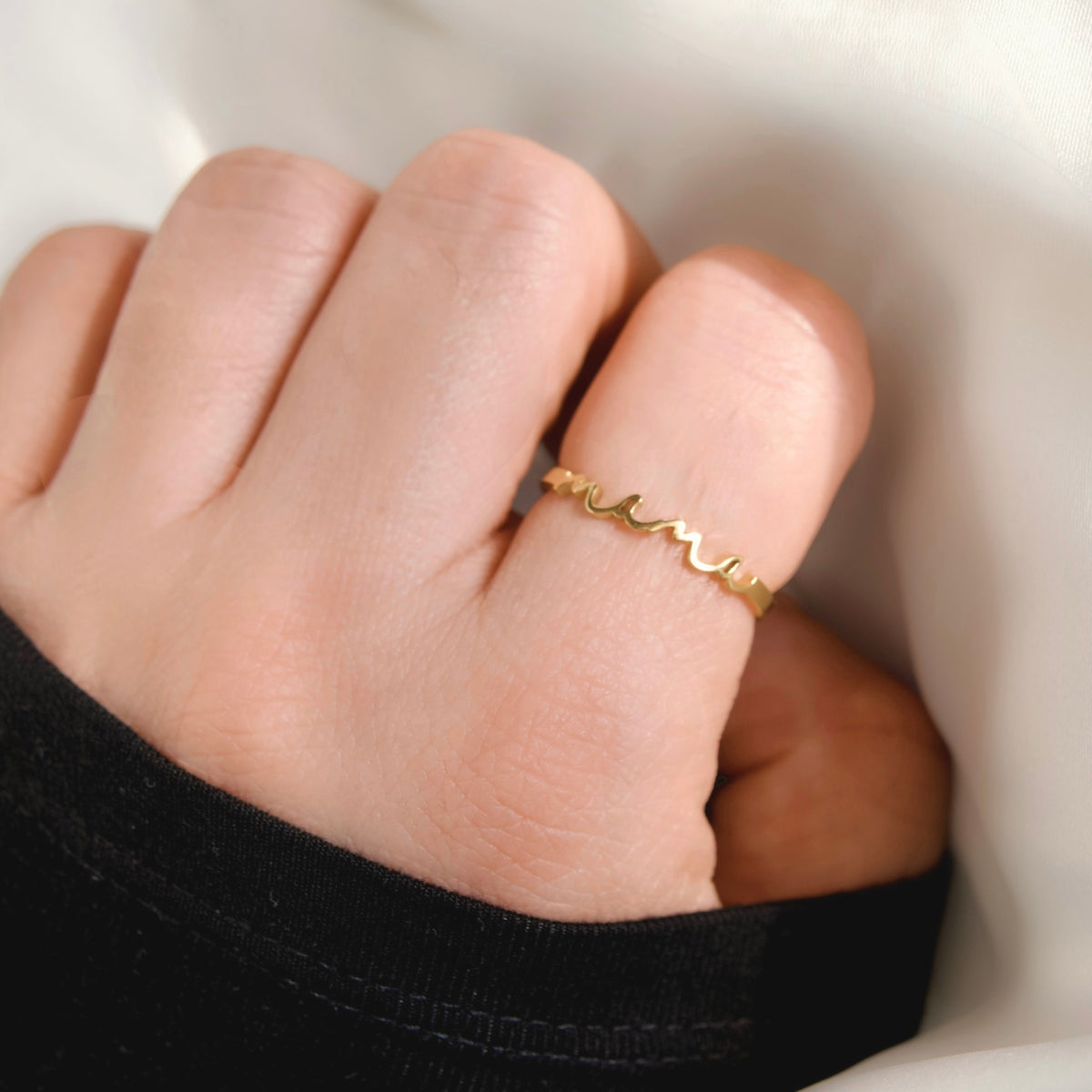 A close-up of a hand wearing a delicate, thin gold ring with a wavy, undulating line design with 'mama' script, set against a soft white background.