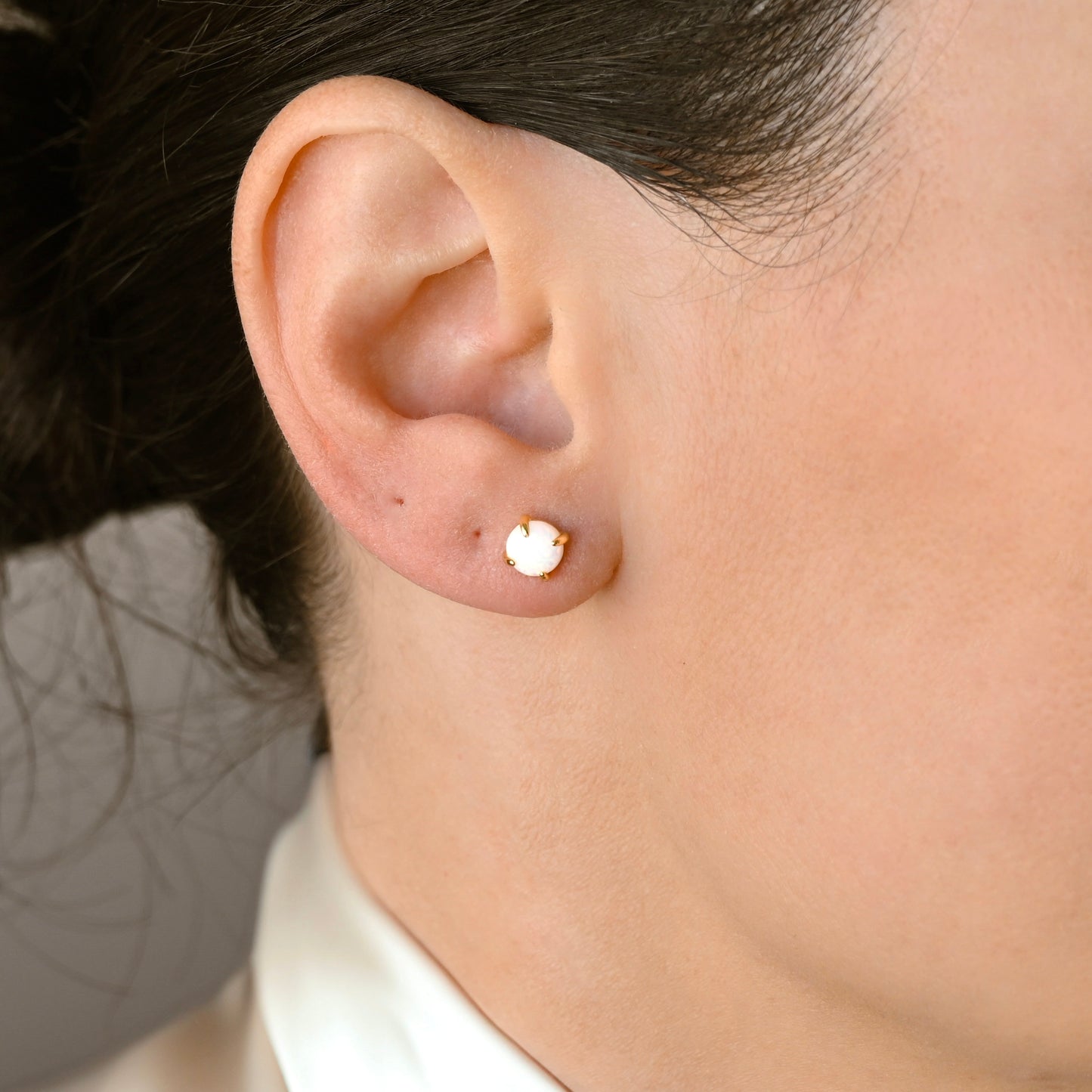 A close-up of a person's ear wearing  small round stud earring with a white opal center and a gold-toned 4-prong setting