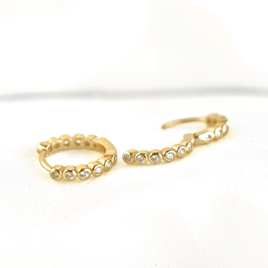 A pair of gold huggie hoop earrings with small clear stones set along the front, displayed on a white textured fabric background.