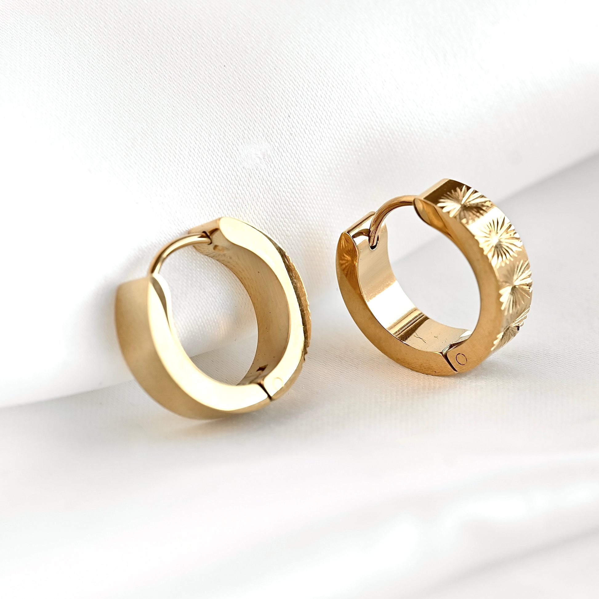 A pair of thick gold huggie earrings one standing upright showing the sun pattern, the other resting against white fabric showing the smooth high polish sides of the earrings.