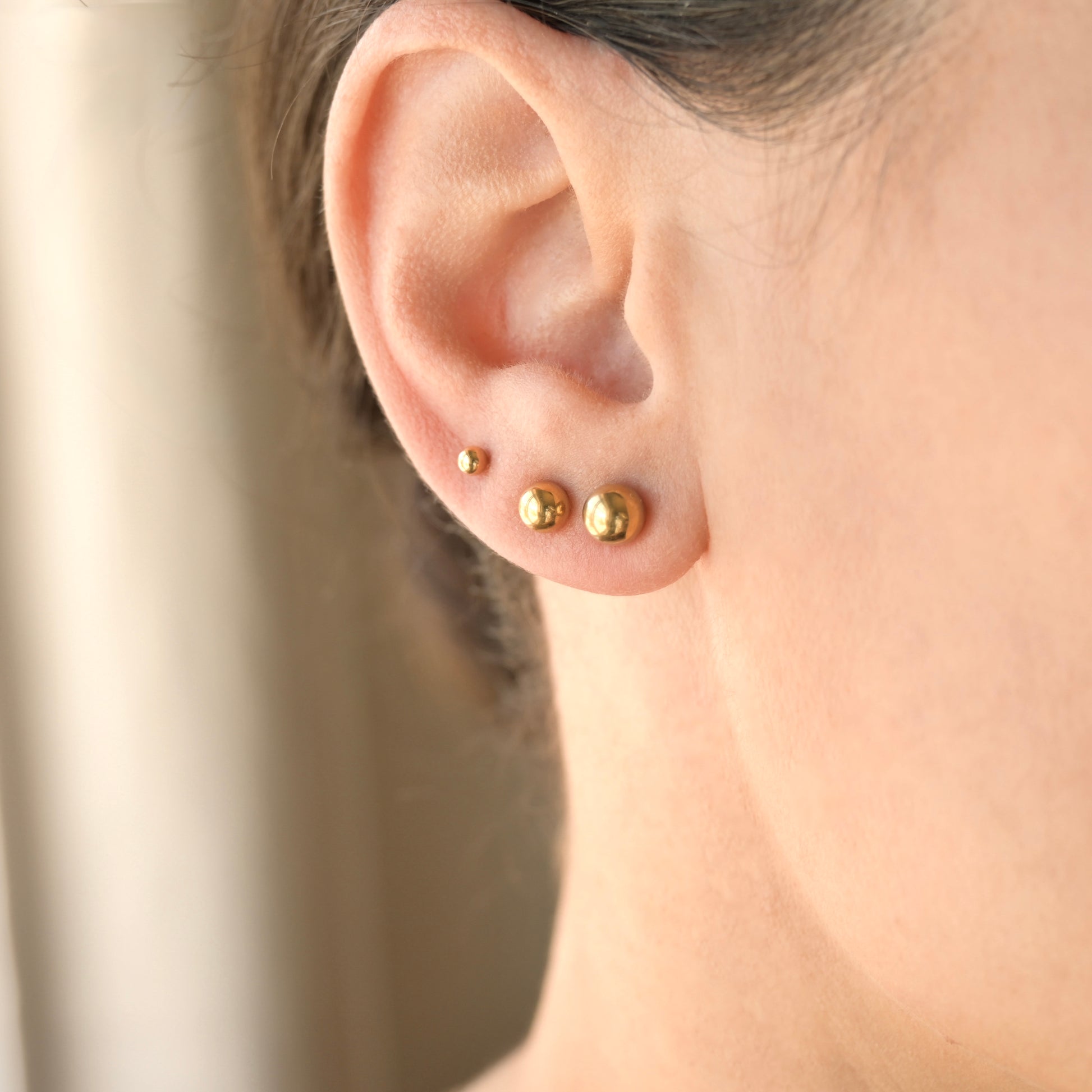 Close-up of an ear wearing gold stud earrings  in three different sizes with a neutral background