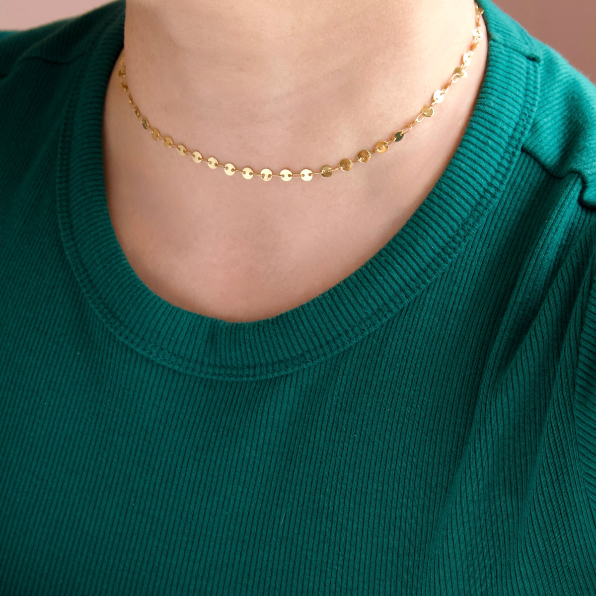 A close-up of a person wearing a delicate gold chain necklace with small flat gold discs, paired with a ribbed teal green top.
