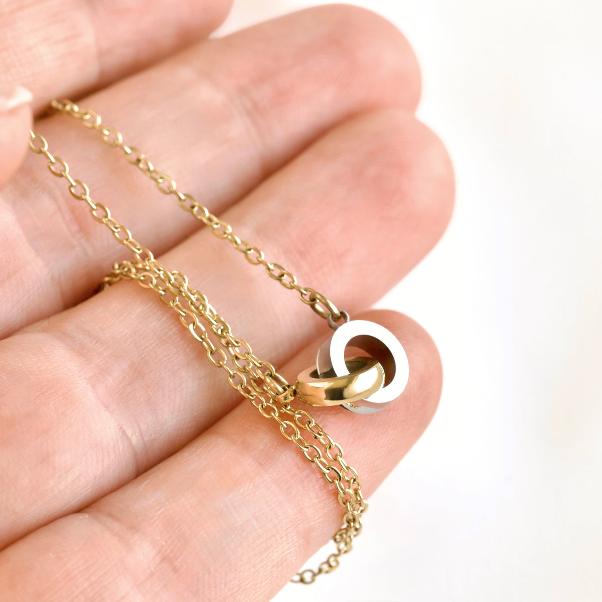 A delicate gold chain necklace with a two-tone interlocking ring pendant, held in the palm of a hand against a bright white background.