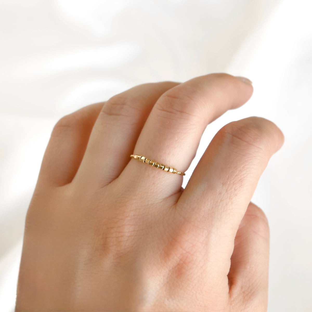 A delicate gold ring with small textured cube-like beads worn on a person's finger, shown against a soft white background.