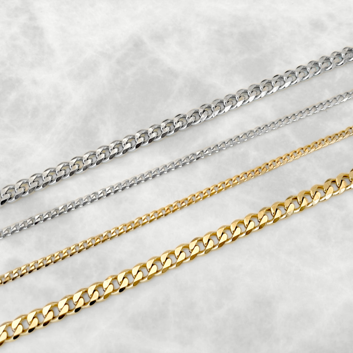 Four metal chain necklaces in silver and gold, arranged diagonally on a light gray textured background.