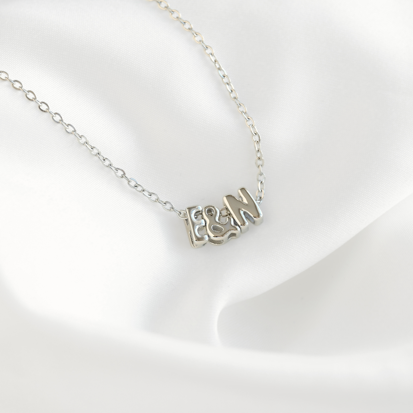 A silver initial necklace featuring the letters 'F', '&', and 'M' in a textured, openwork design, displayed on a soft white satin fabric background.