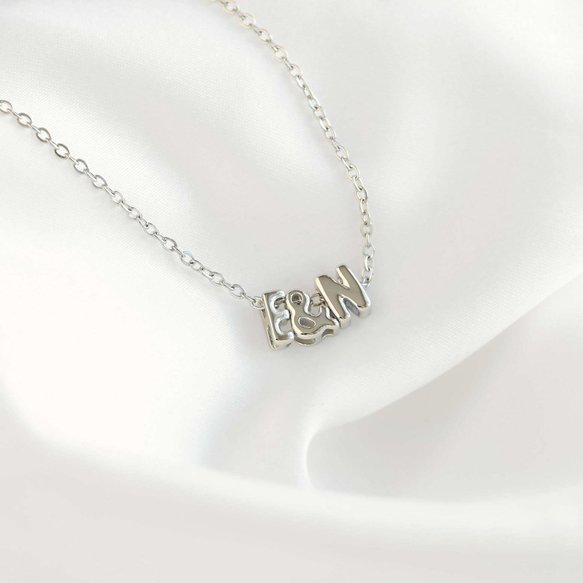 A silver initial necklace featuring the letters 'F', '&', and 'M' in a textured, openwork design, displayed on a soft white satin fabric background.