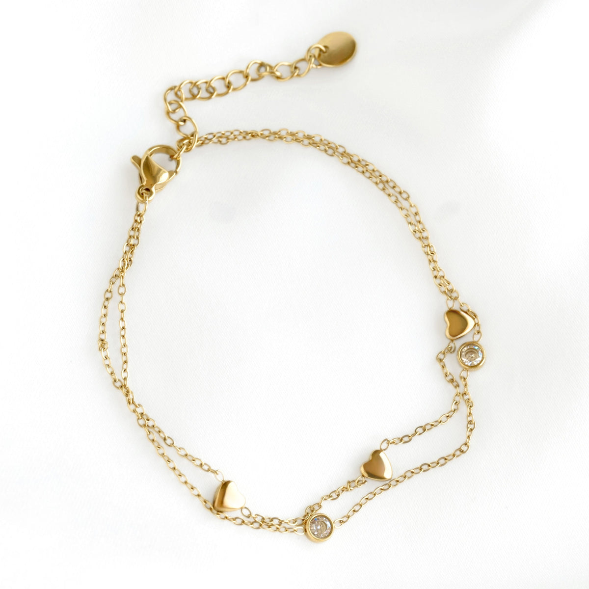 A delicate gold chain bracelet featuring small heart charms and two round bezel  stone accents, displayed on a clean white background.