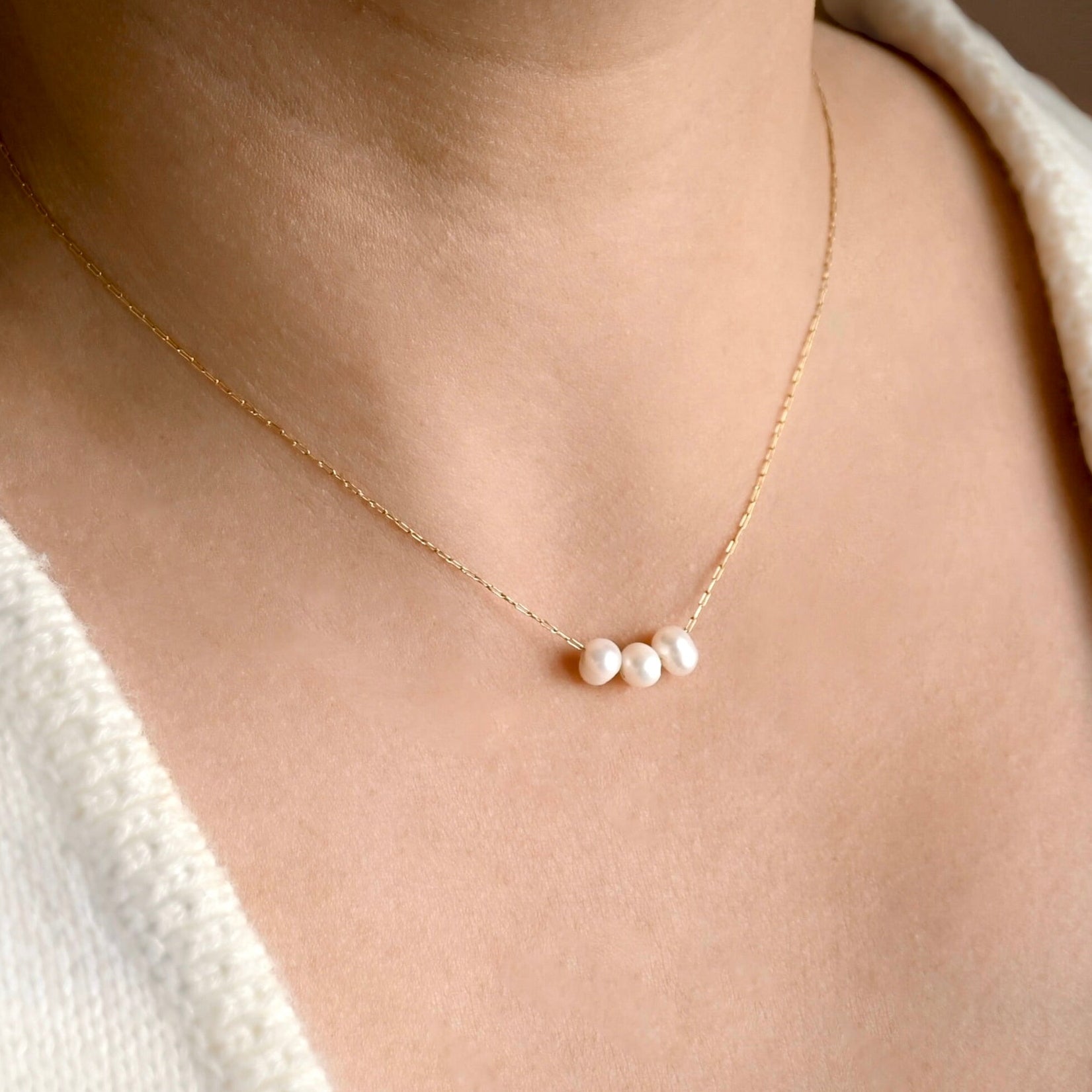 A delicate gold chain necklace with three small white pearls, worn on a model's neck over a cream-colored knit top.