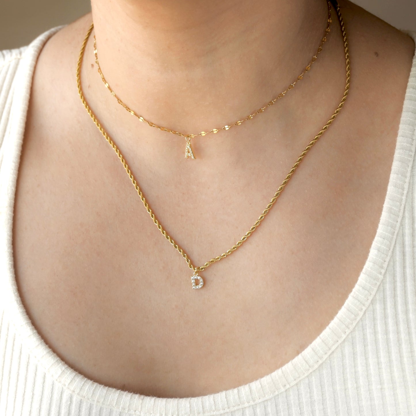 Front view two gold necklaces layered, one shorter mariner chain with letter 'A', and one longer rope chain with letter 'D' on a person's neck wearing a white top