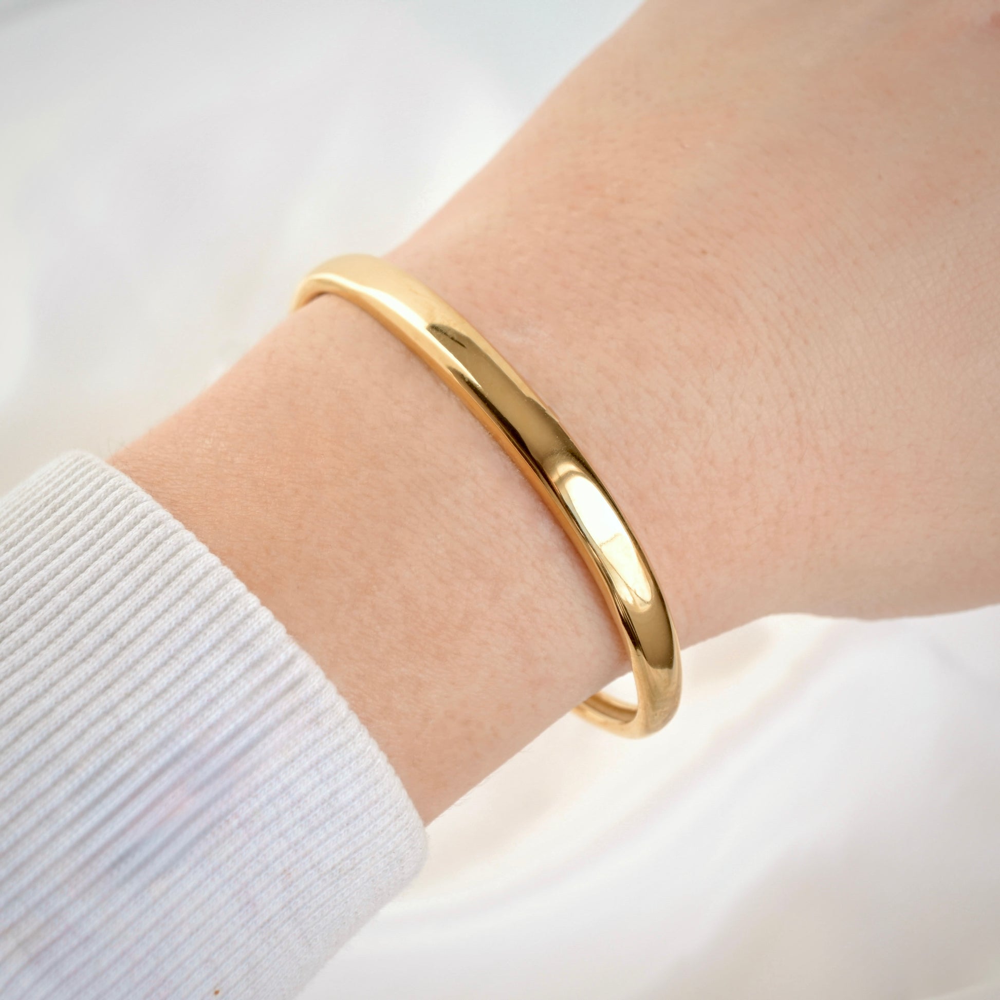 A polished gold cuff bracelet worn on a person's wrist, shown against a soft white background with a white ribbed sleeve visible.