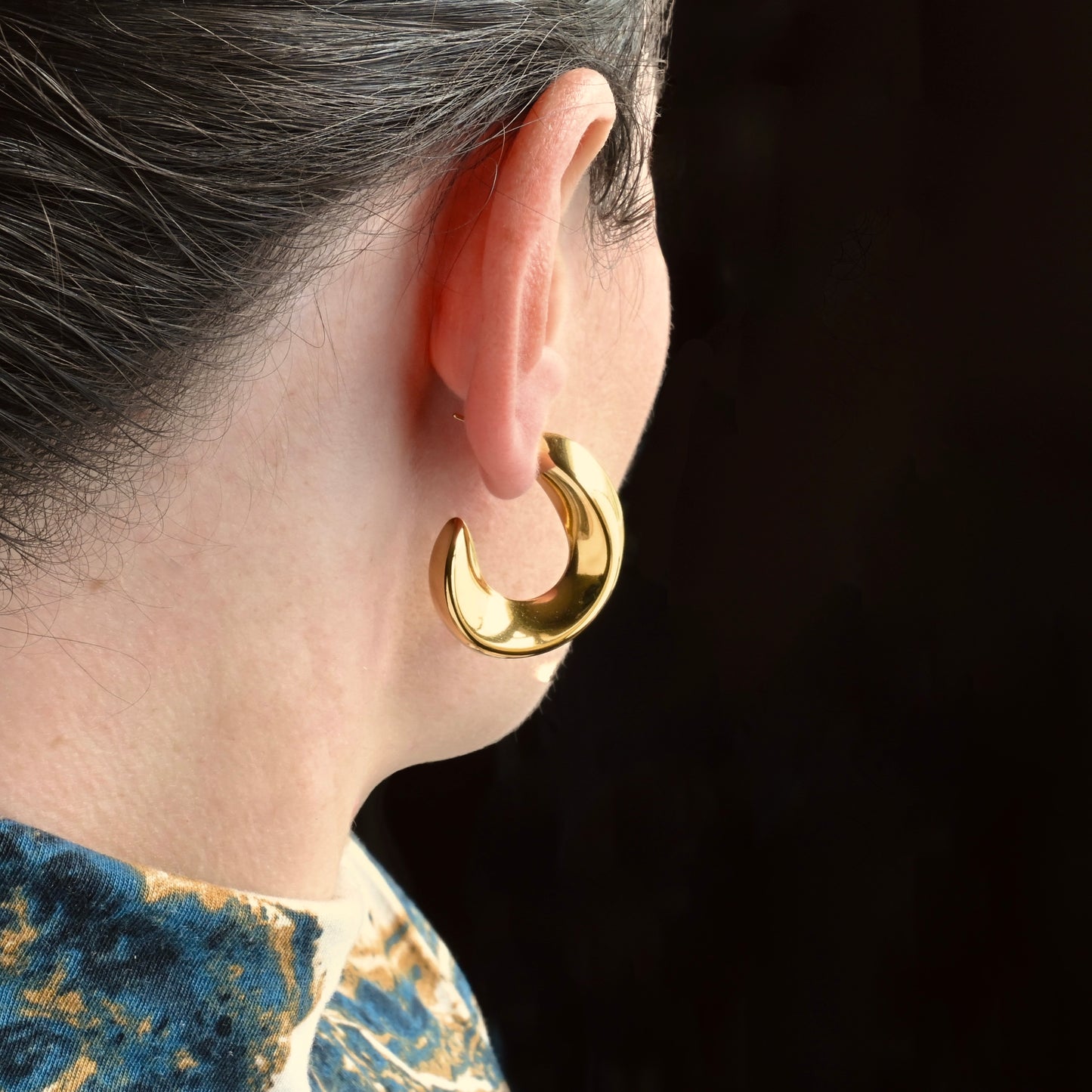 Organic sculptural C hoop earrings worn by a person against a dark background