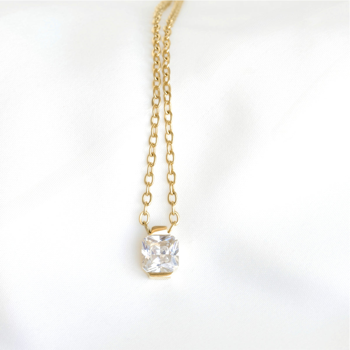 A dainty gold chain necklace with a small square clear gemstone pendant, displayed against a soft white fabric background.