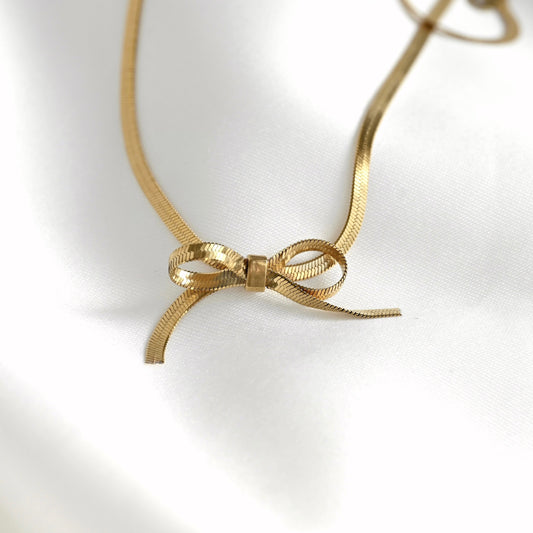 A gold-colored snake chain necklace featuring a delicate bow-shaped pendant, displayed on a soft white fabric background.
