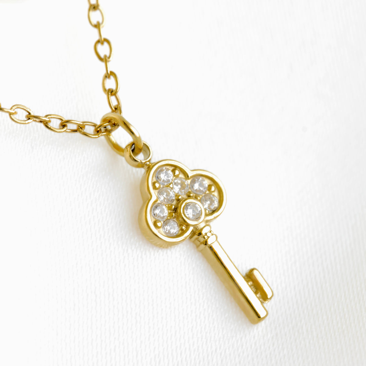 A gold key pendant necklace on a delicate gold chain, featuring a flower-shaped head paved with clear crystals, set against a clean white background.