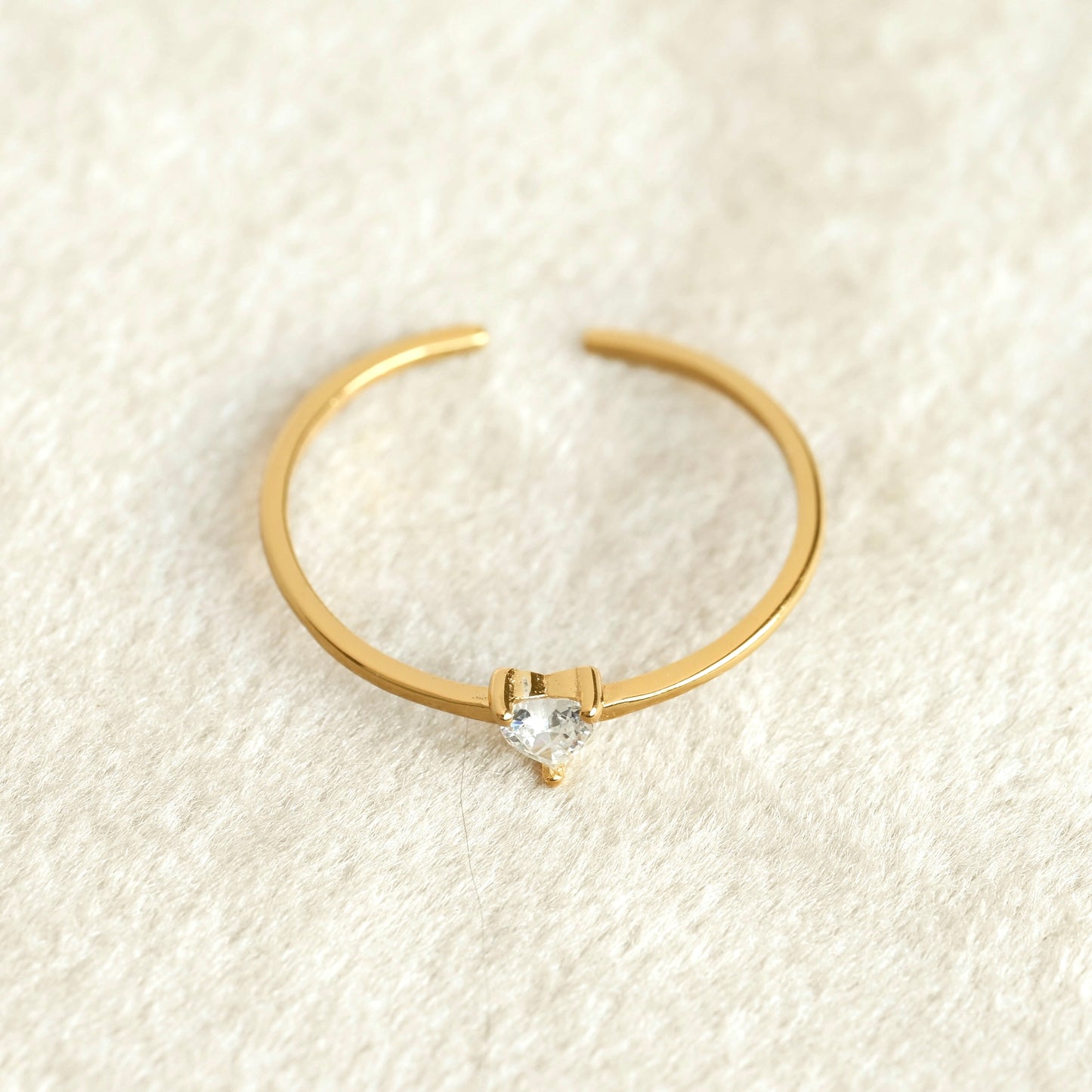 Top view of Aa delicate gold open ring with a small clear heart-shaped gemstone in the center, resting on a textured cream-colored fabric background.