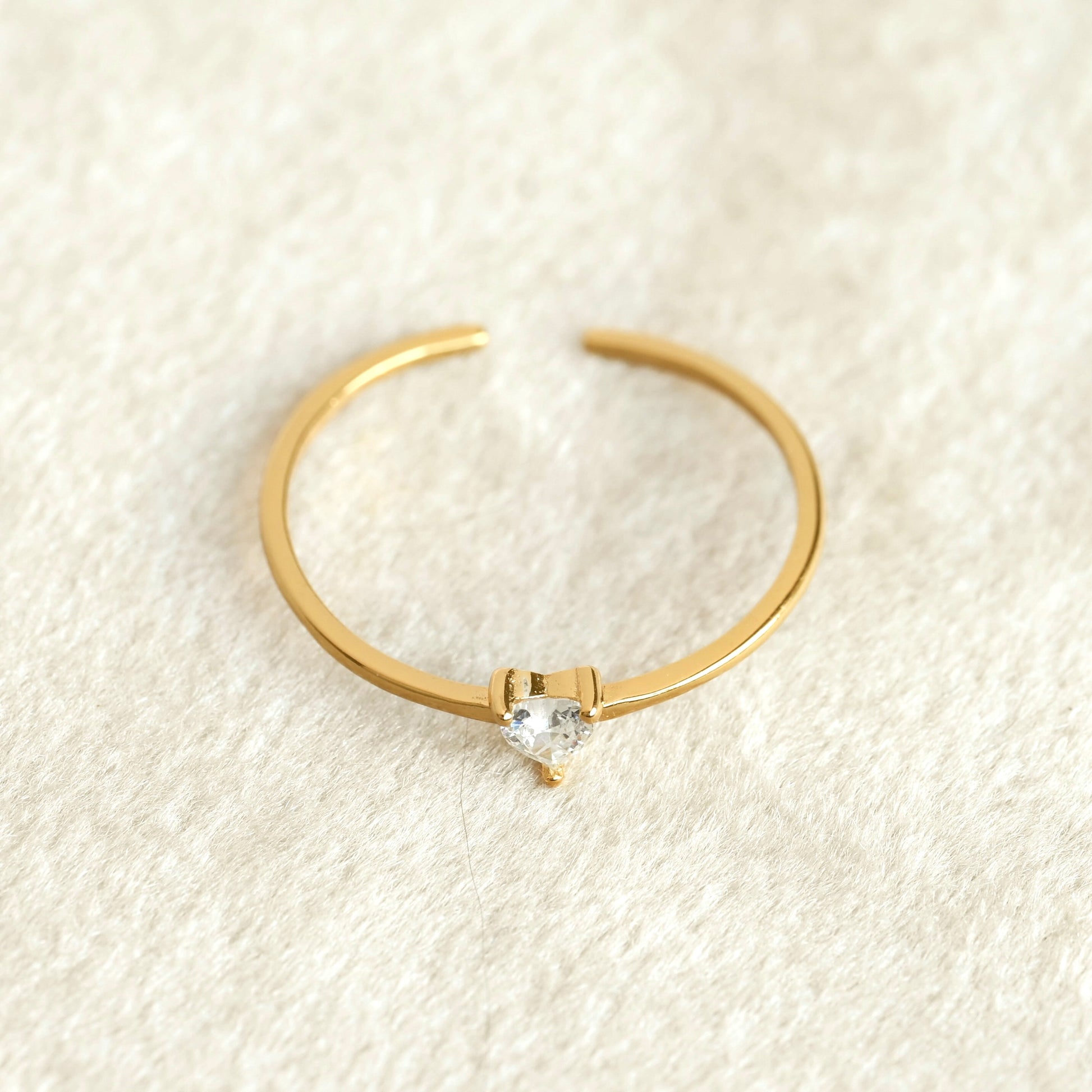 Top view of Aa delicate gold open ring with a small clear heart-shaped gemstone in the center, resting on a textured cream-colored fabric background.