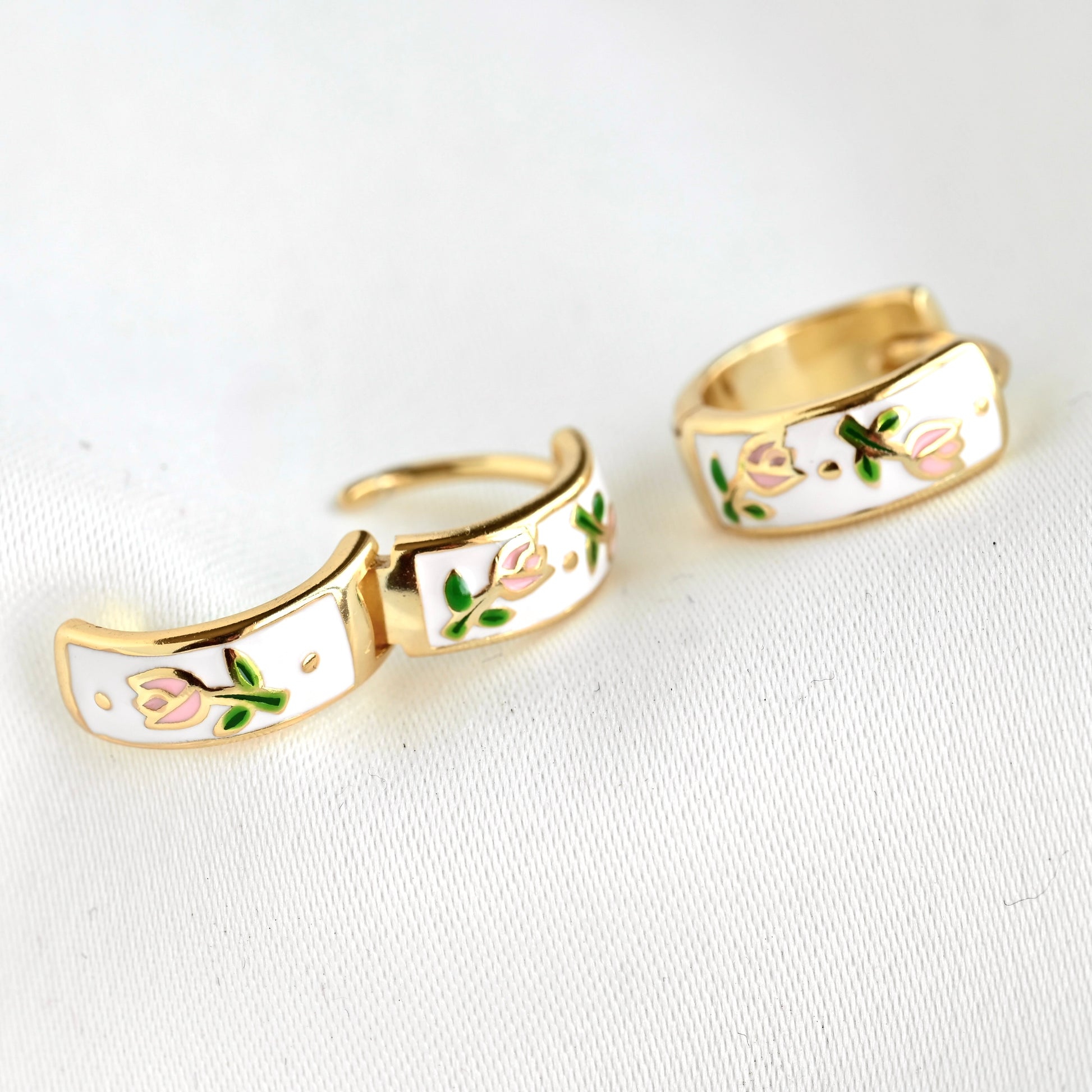 A pair of gold-toned hoop earrings with white enamel inlay, decorated with pink tulip flowers and green leaves, displayed on a textured white fabric background.