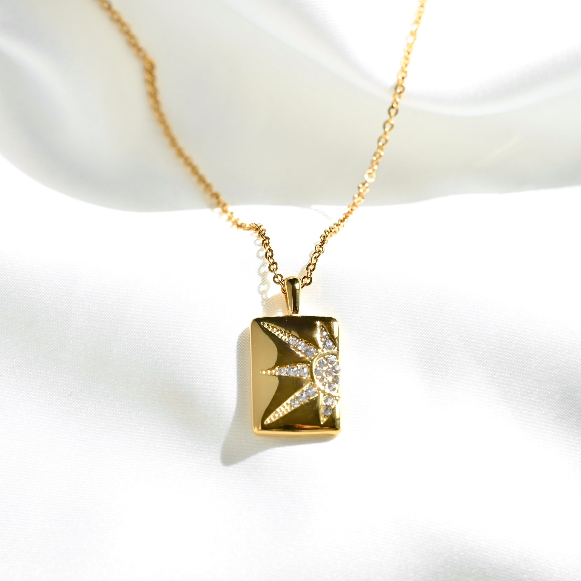 A gold pendant necklace featuring a rectangular gold charm with a starburst design set with clear crystals, displayed on a white satin fabric background.