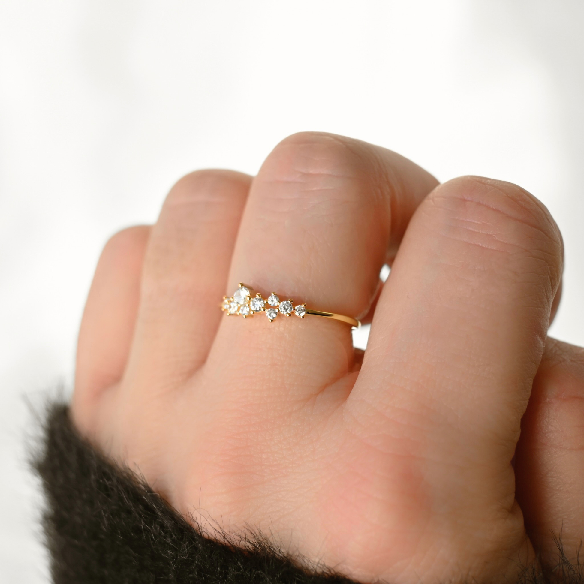A delicate gold ring with small clear gemstones set across the middle finger of a hand, shown in a close-up shot against a bright, soft white background.