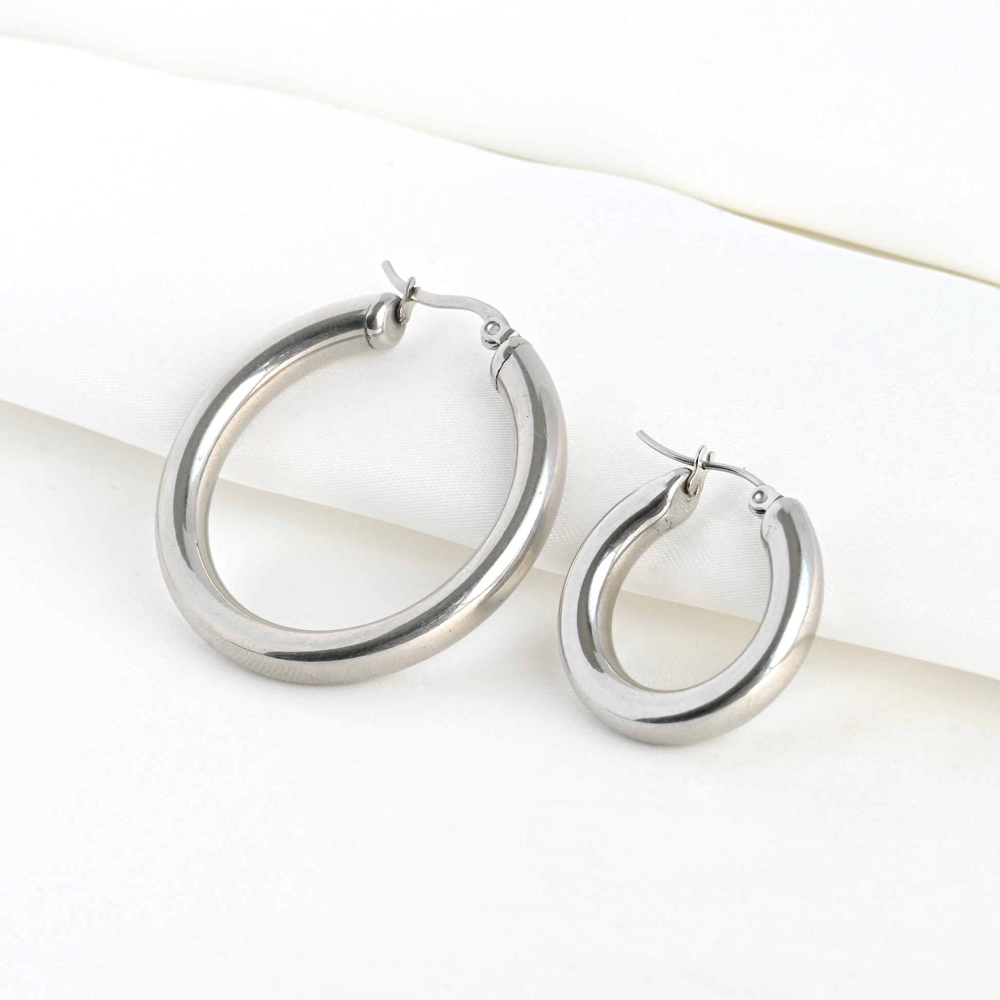 One small and one medium sized thick silver hoop earrings on a white satin fabric