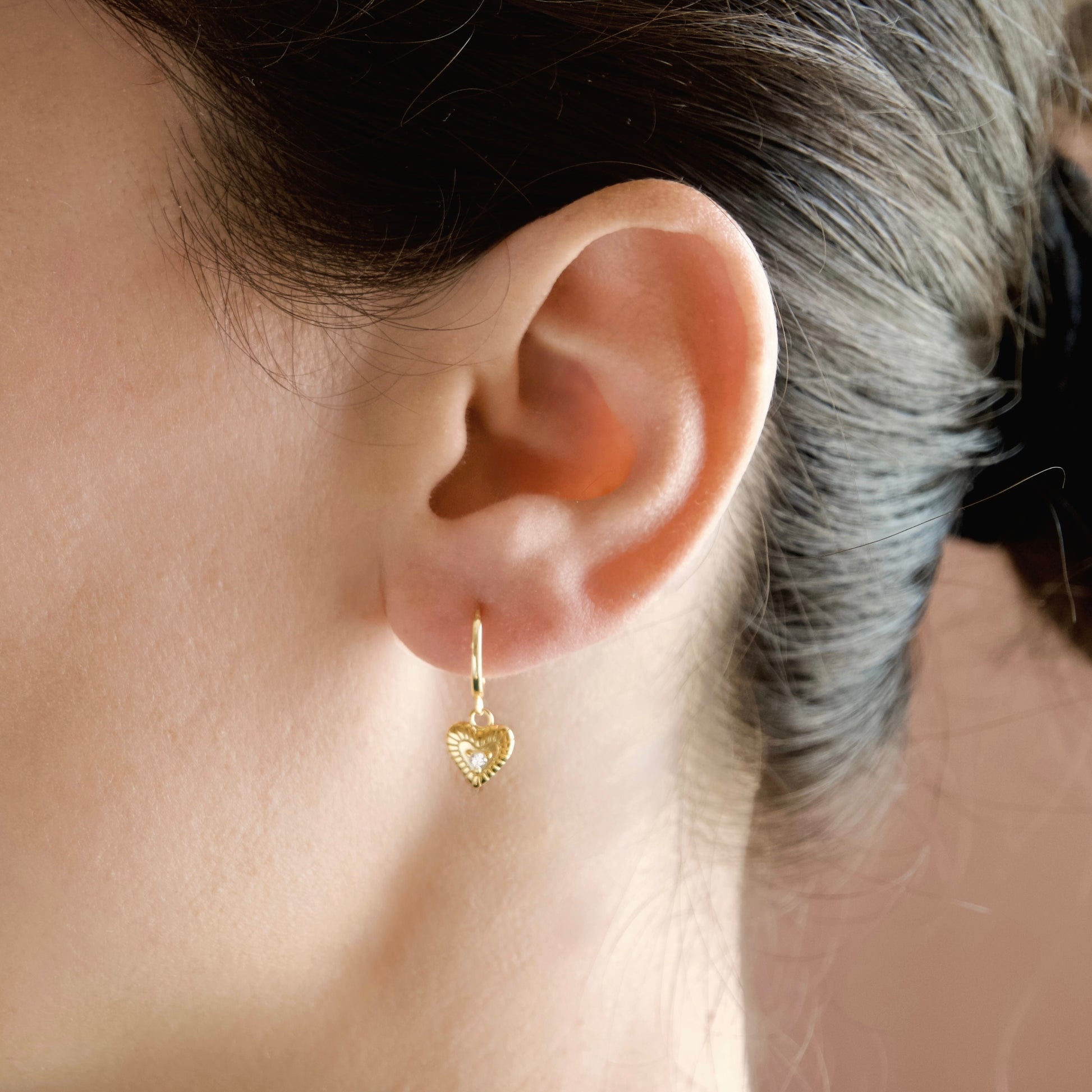 A close-up of a person's ear wearing a small gold huggie hoop earring with a dangling textured heart charm.