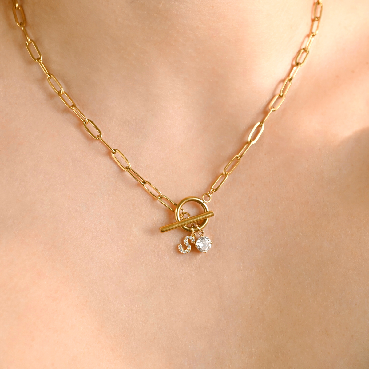A close-up of a gold paperclip chain necklace with a toggle clasp and small dangling clear stones, worn on a person's neck.