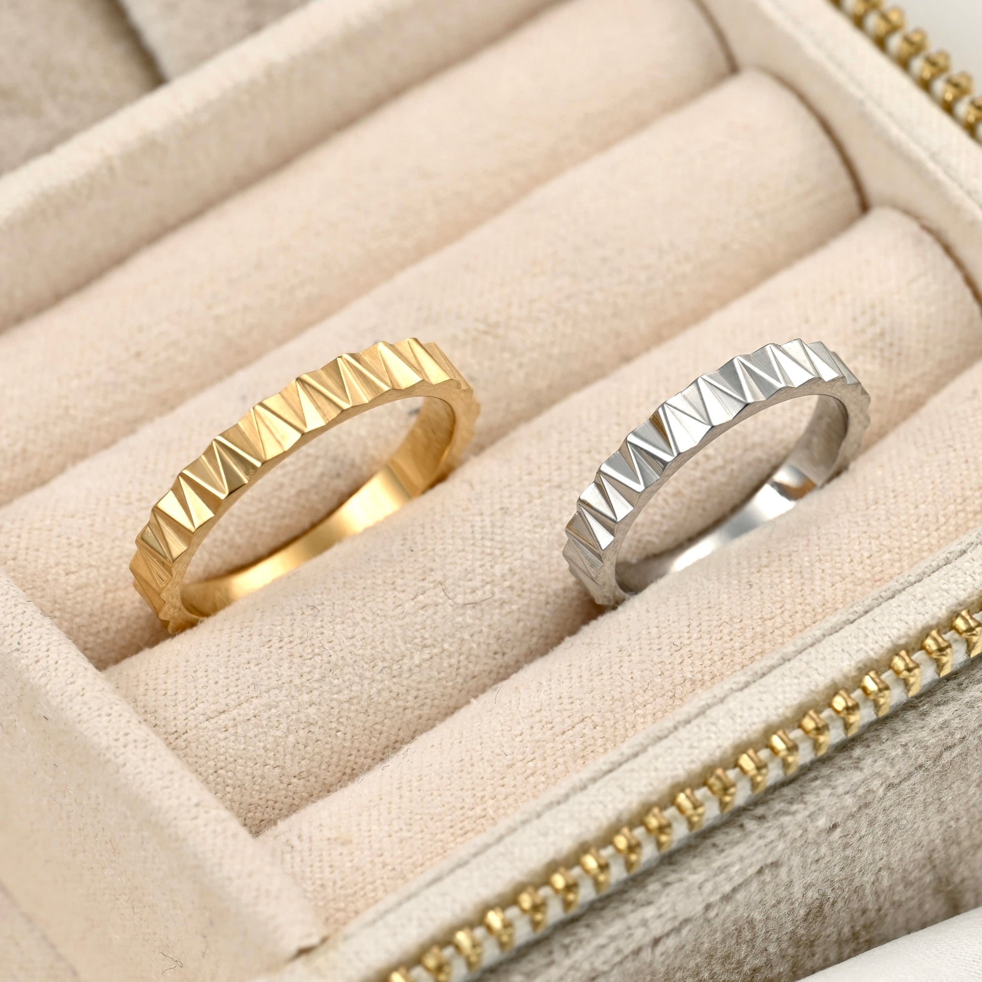 Two geometric pyramid-stacked rings, one gold and one silver, displayed in a beige fabric ring box.