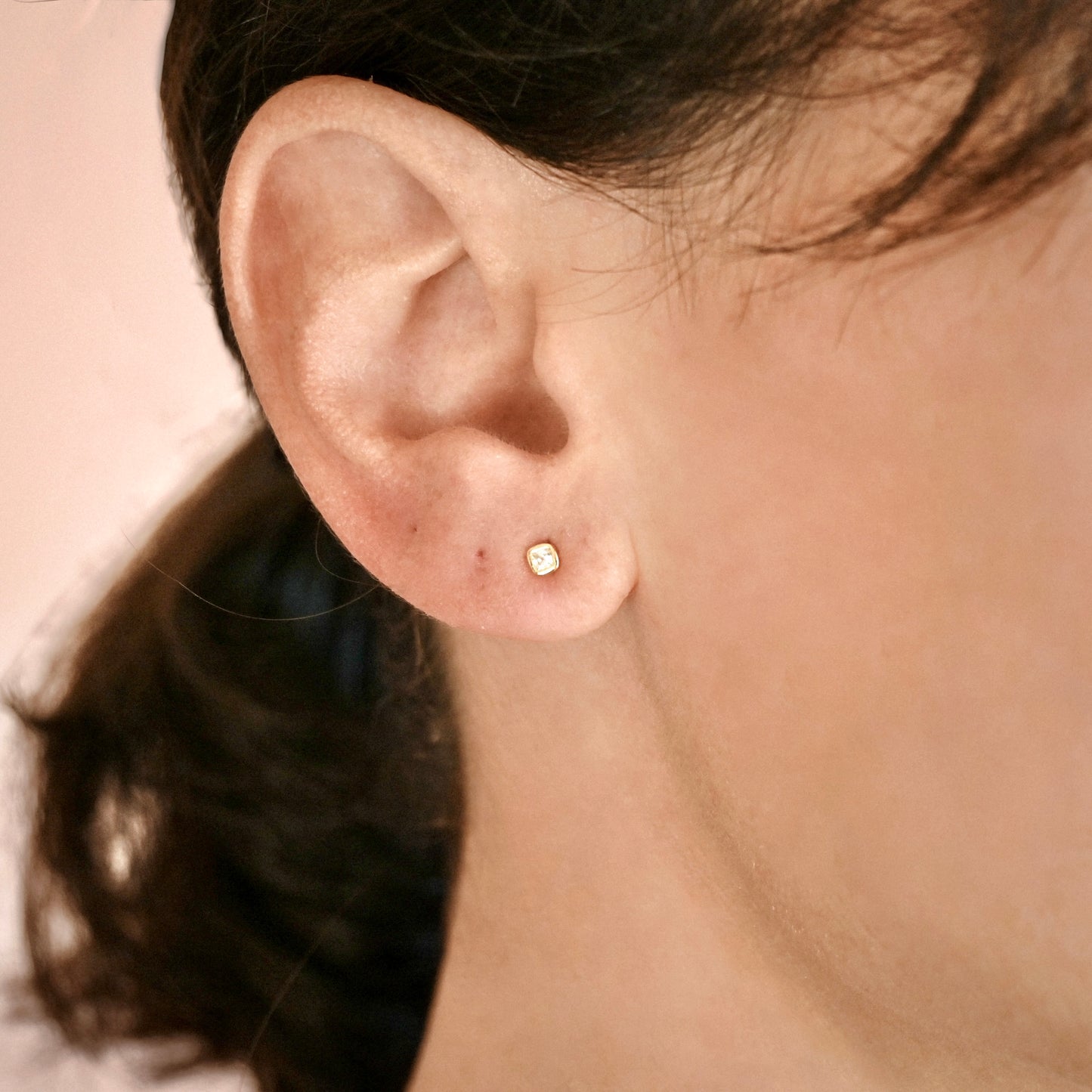 A close-up of a person's ear wearing a small square gold stud earring.