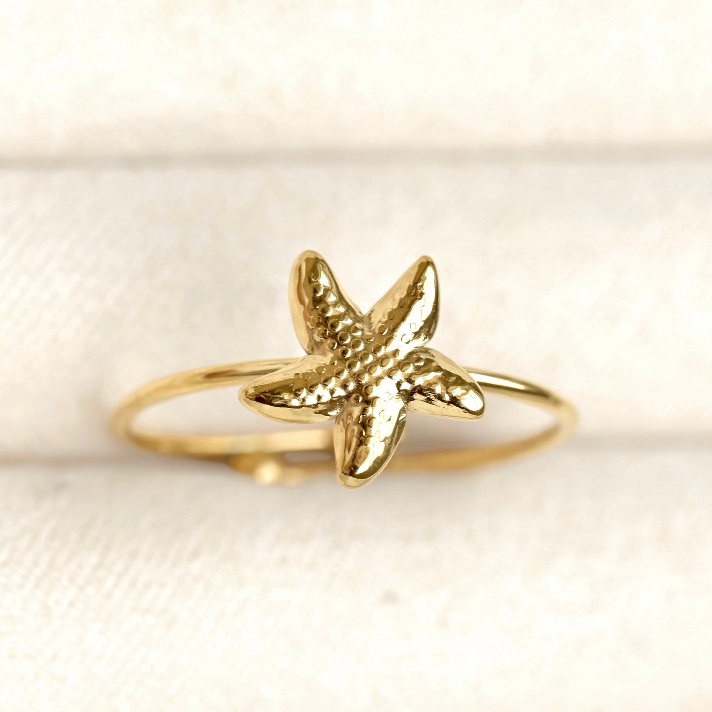 A gold starfish ring with a textured, hammered finish on a polished band, displayed on a soft beige fabric background.