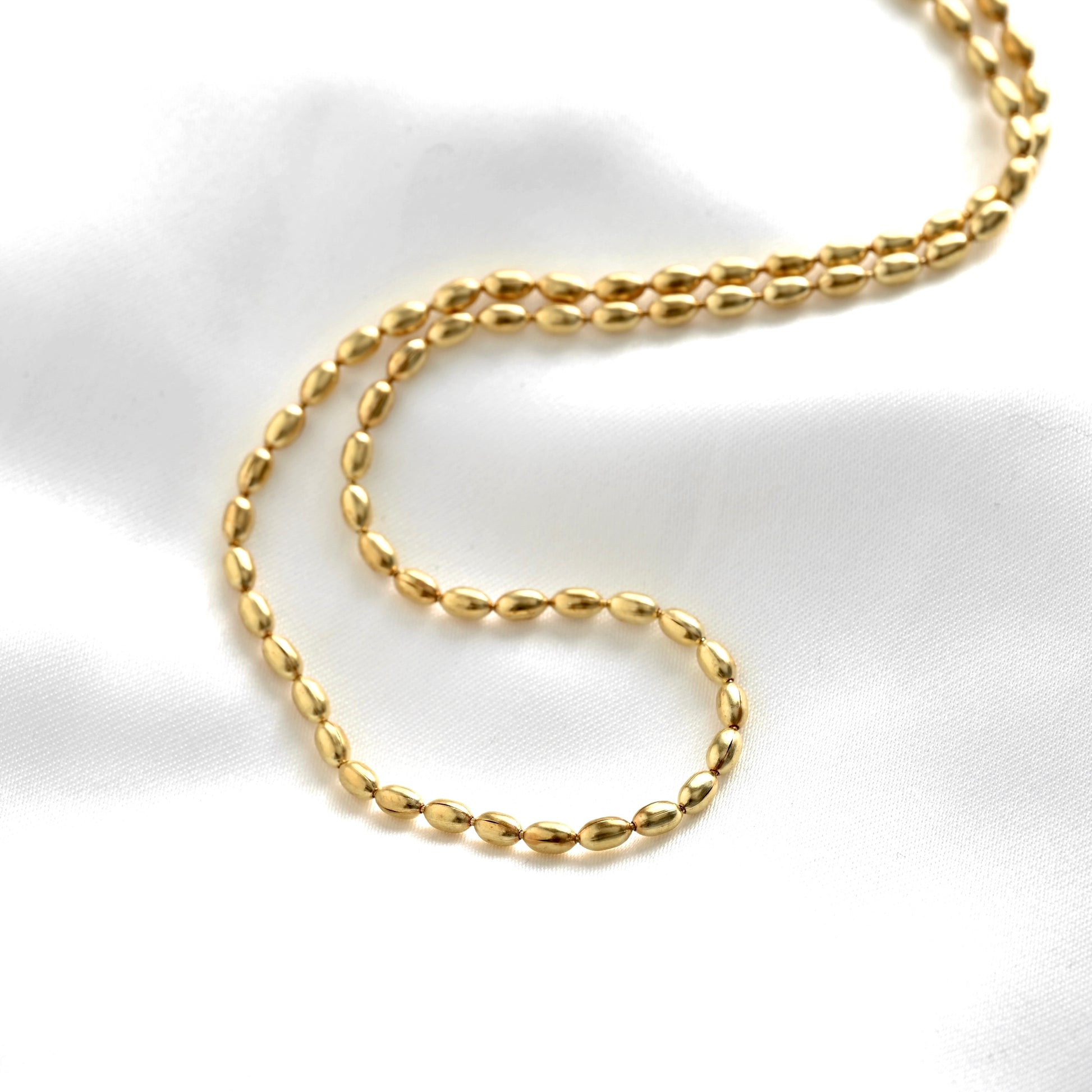 A close-up of a gold chain necklace with a textured, polished oval beaded design, arranged in a loop on a white satin fabric background.