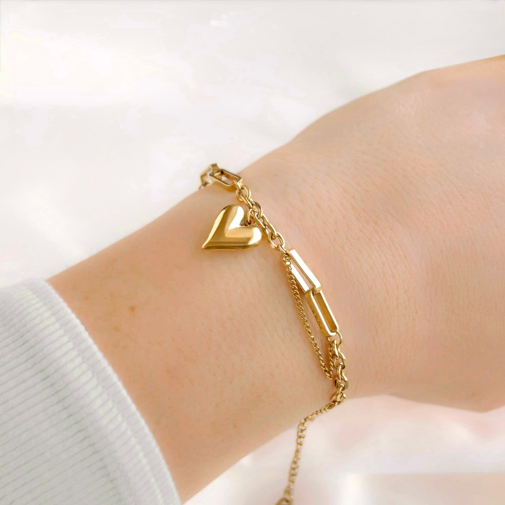 A delicate gold bracelet with a heart charm and a polished paperclip link detail, shown worn on a person's wrist.