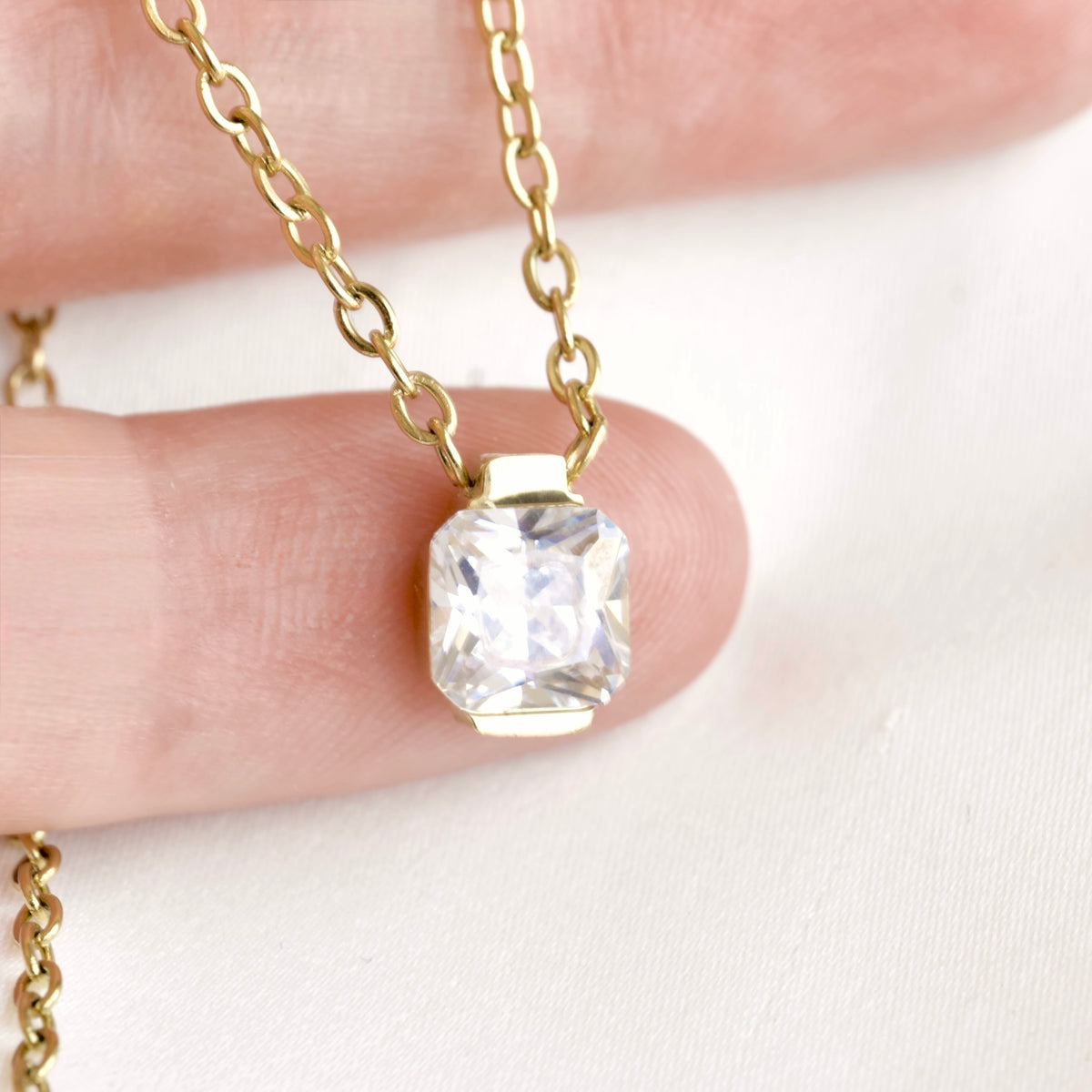 A close-up of a gold chain necklace with a cushion-cut clear gemstone pendant, held between two fingers against a soft white background.