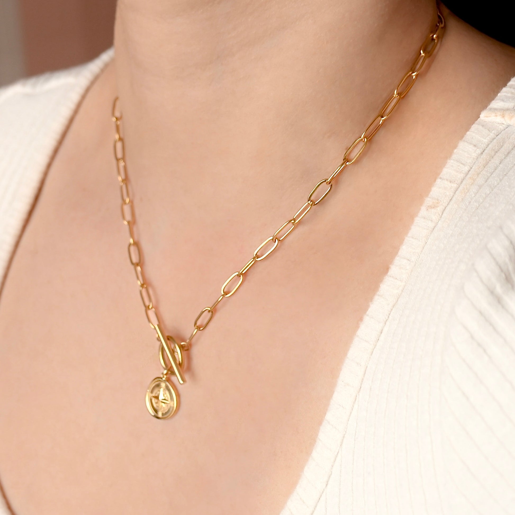 A close-up of a gold chain necklace with a toggle clasp and a small oval pendant, worn on a model's neck over a white ribbed top.